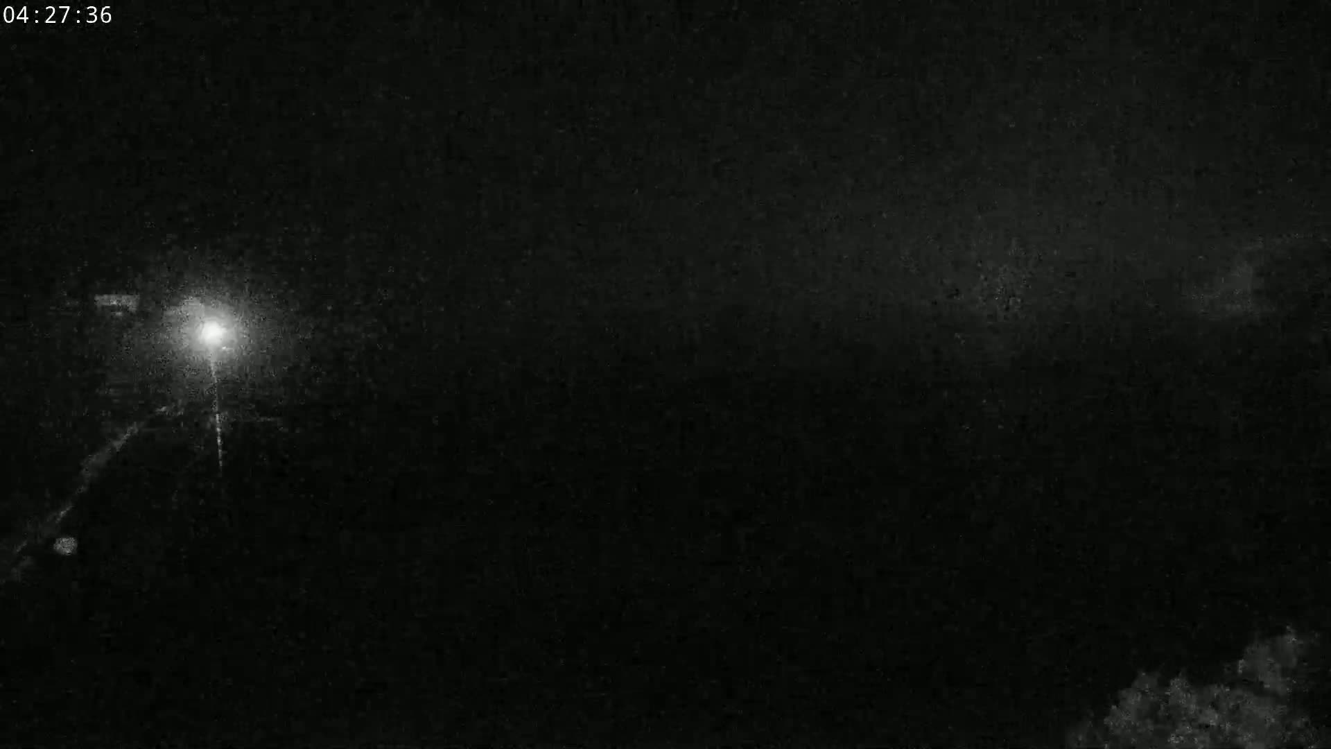 A nighttime, low-light image shows a bright light source in the distance with faint light trails suggesting vehicle headlights on a dark road.