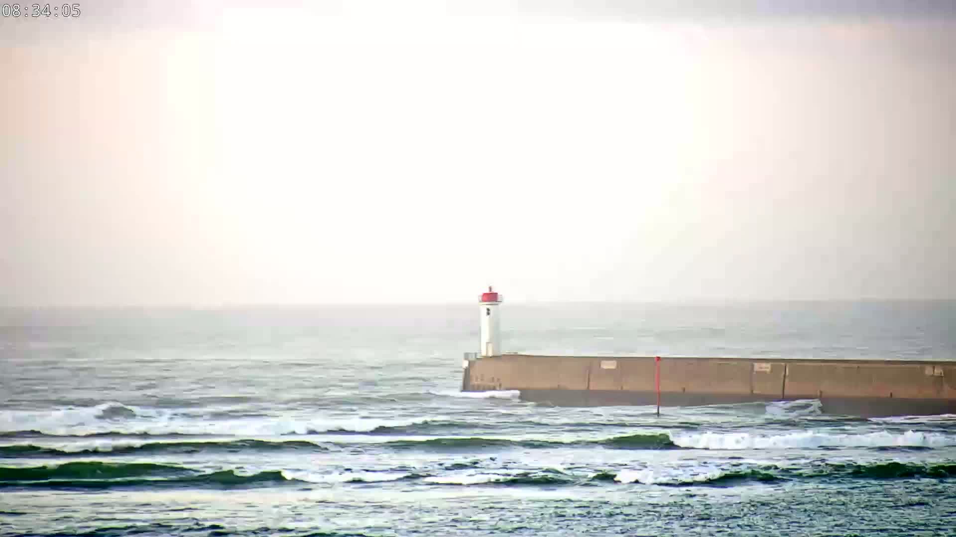 A small lighthouse sits atop a breakwater in a choppy sea under a hazy sky.