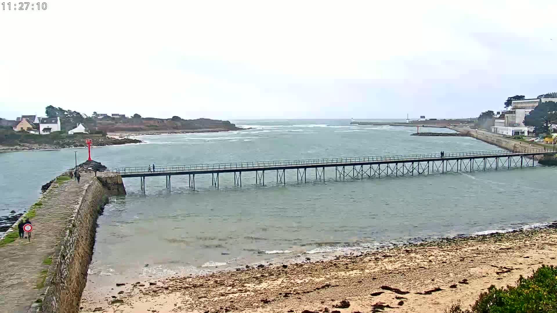 A long pier stretches across calm, gray water, connecting a stone jetty to a distant shoreline under an overcast sky.