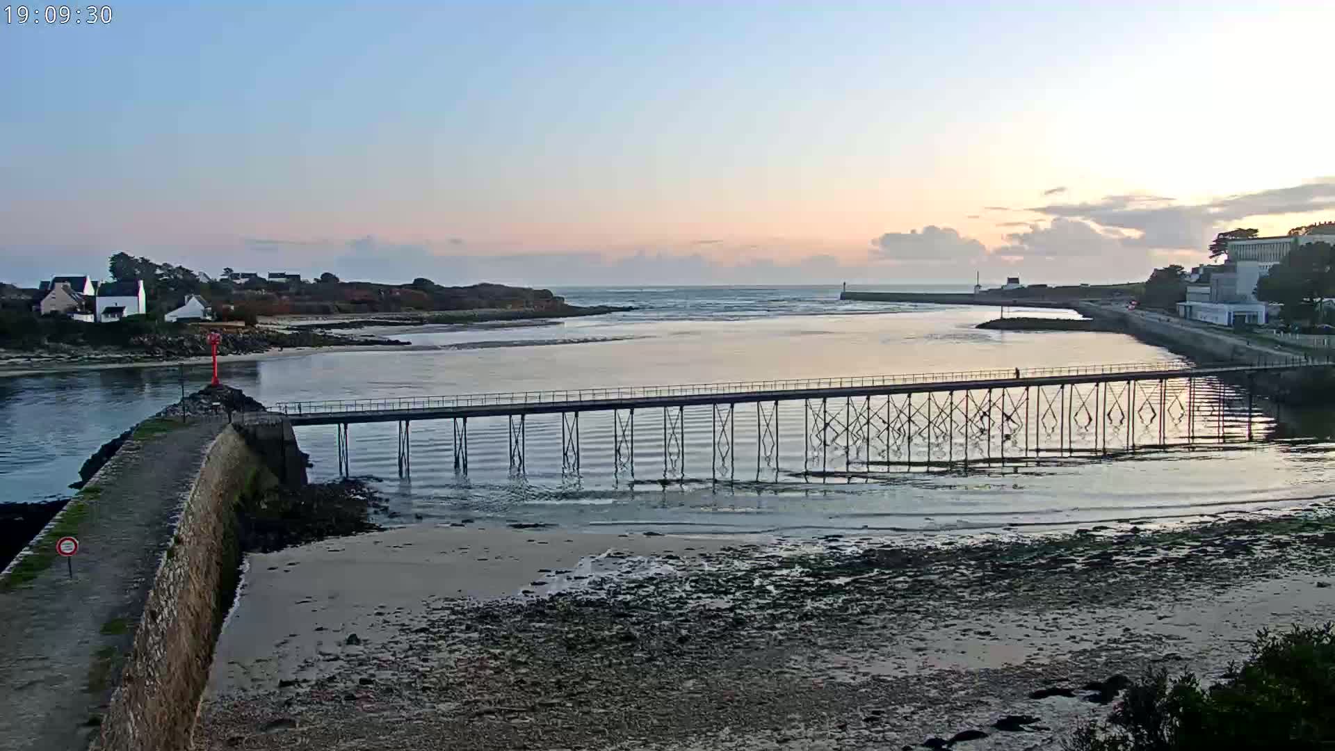A long pier extends over calm water at sunset, connecting a stone jetty to a shoreline with buildings and low hills in the background.