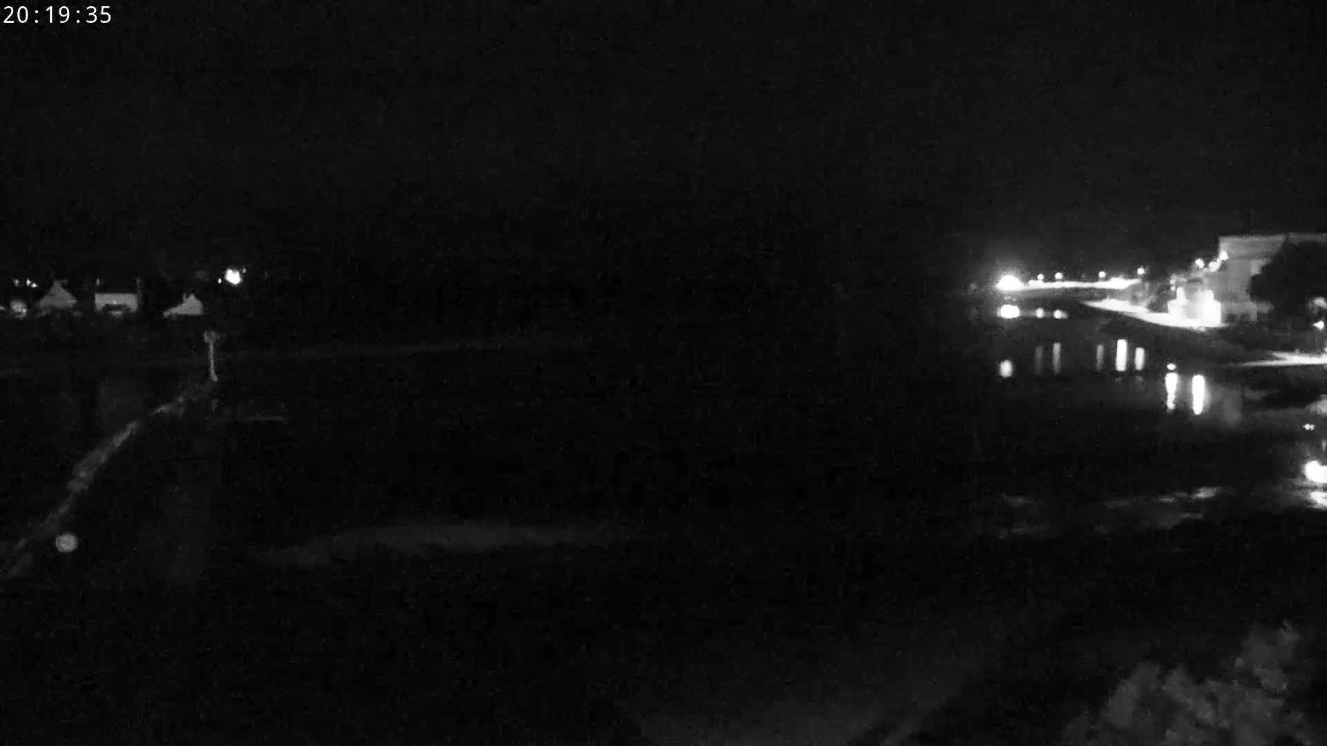 A nighttime, low-light view shows a dark body of water reflecting faint lights from buildings and structures on the opposite shore.