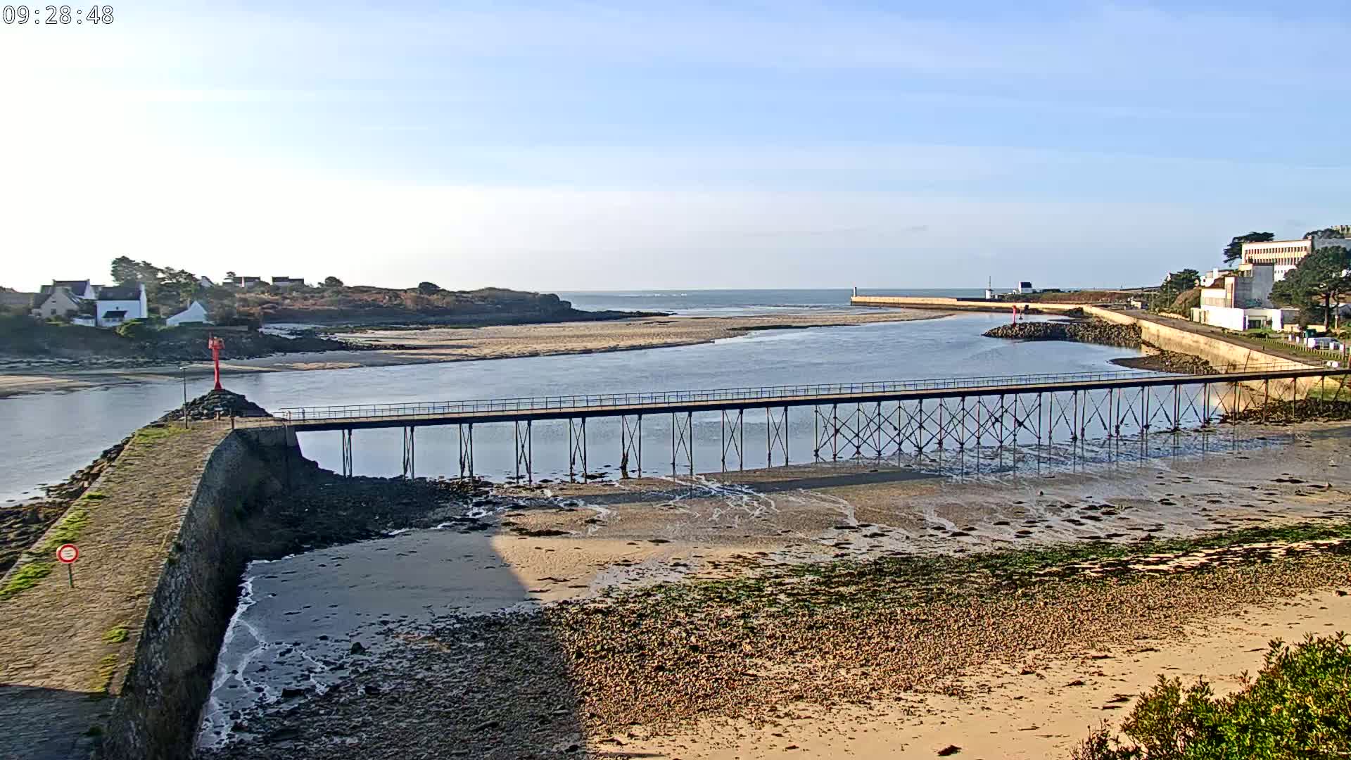 A long pier extends across a calm body of water at low tide, connecting a rocky shoreline to a small village under a clear, sunny sky.