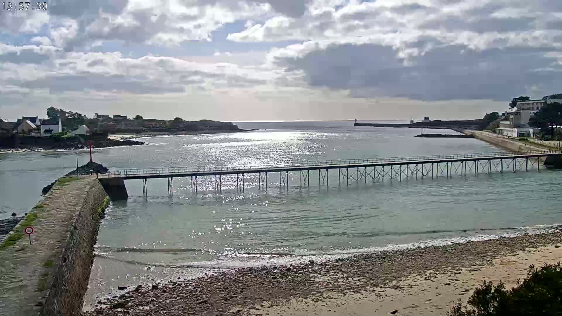 A long pier extends over calm, sparkling water toward a breakwater, under a partly cloudy sky.
