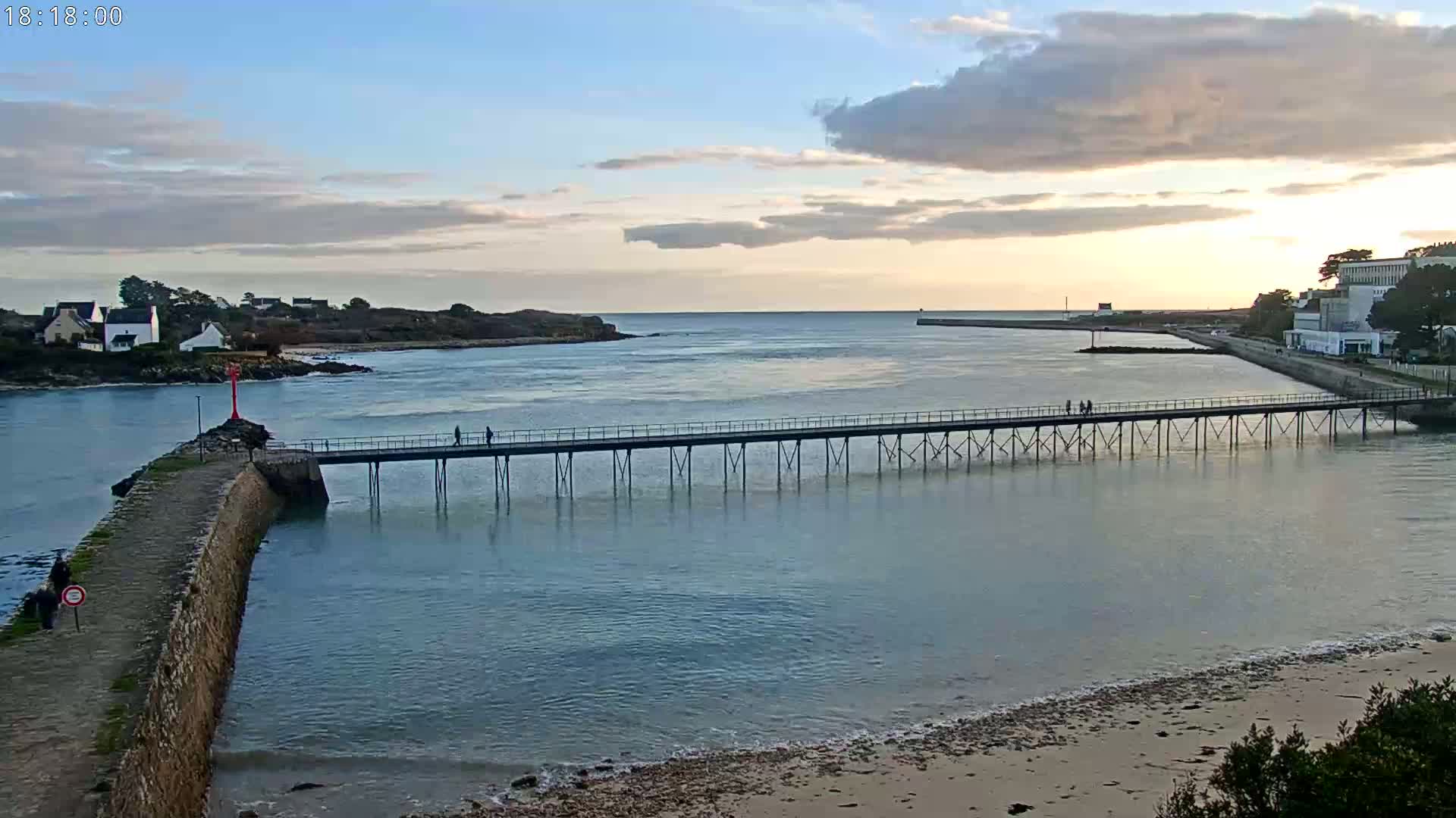 A long pier stretches across calm water under a partly cloudy sunset sky, connecting a stone jetty to a distant shoreline with buildings.