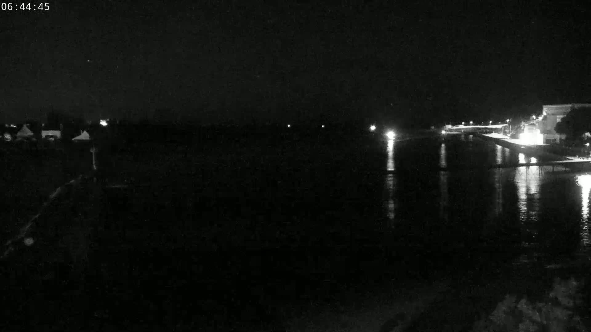 A nighttime view of a calm body of water reflecting lights from buildings and structures on the opposite shore under a clear, dark sky.