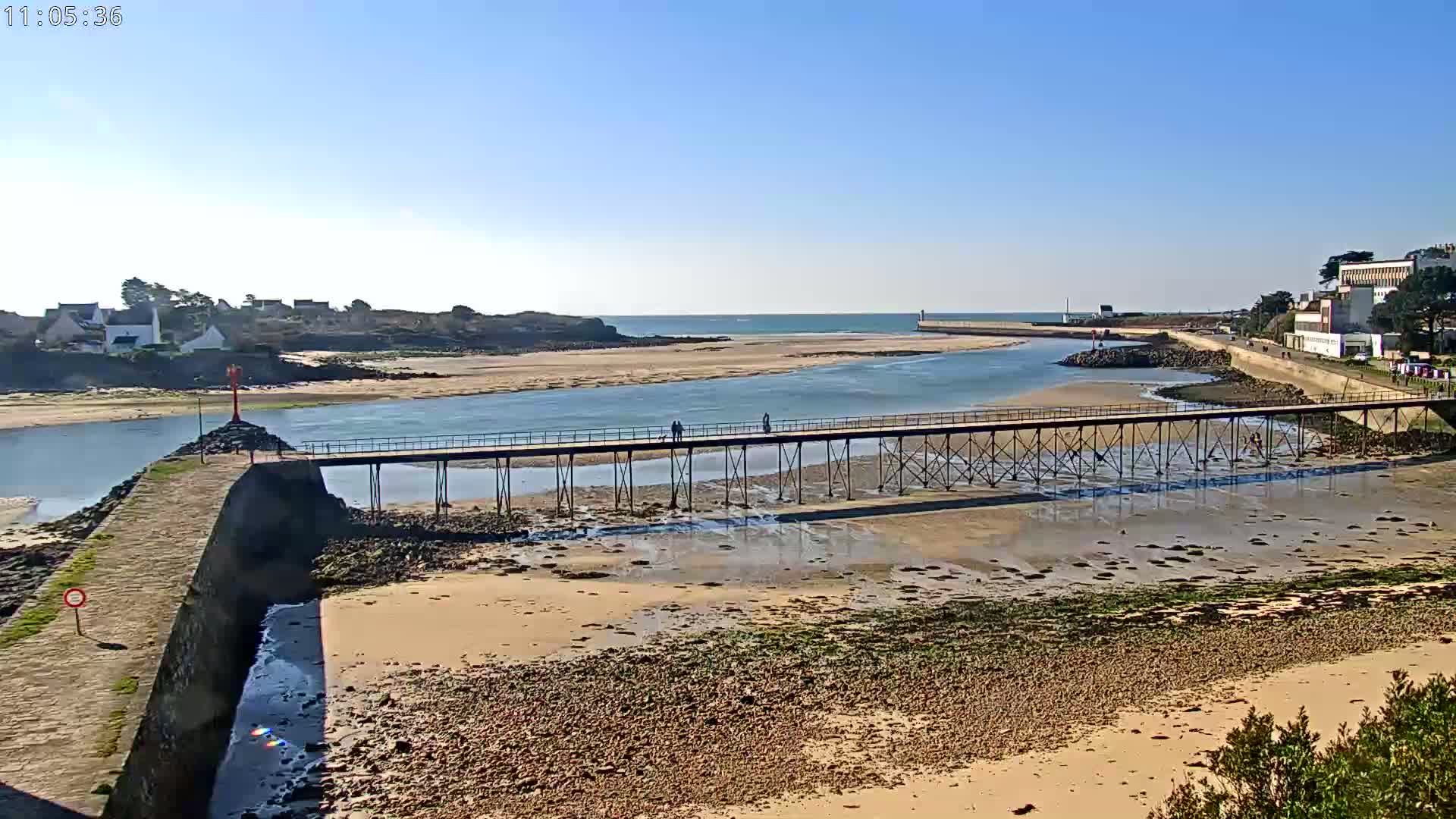 A long pier stretches across a calm waterway at low tide, connecting a small village to a larger town under a clear blue sky.