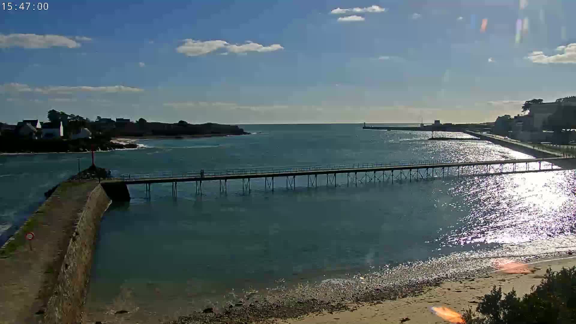A long pier extends over a calm, sunlit body of water toward a distant shoreline under a mostly clear, bright sky.