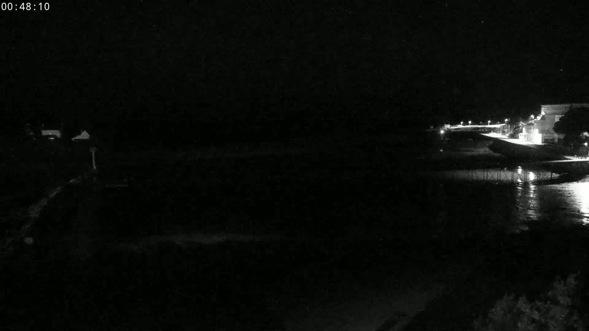 A nighttime, low-light view of a calm body of water near a dimly lit shoreline with buildings in the distance.