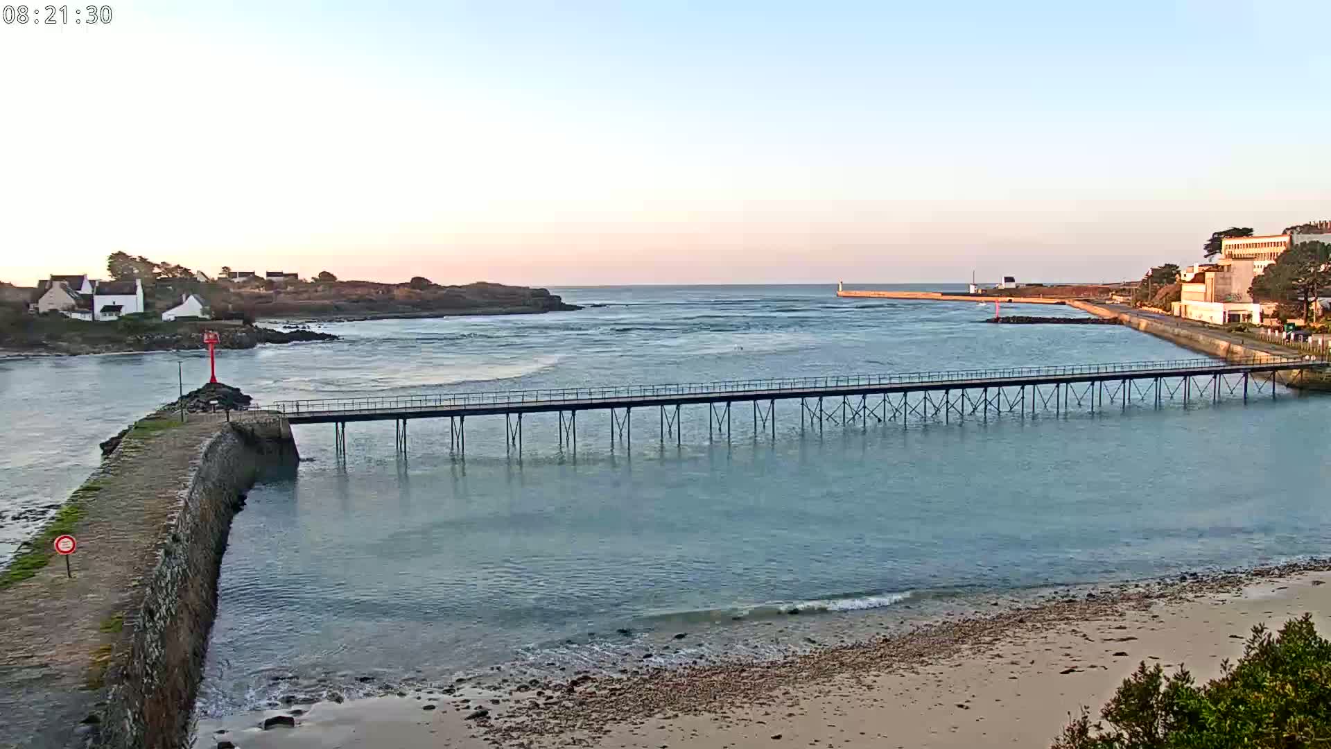 A long pier stretches across calm, light-blue water toward a distant shore under a pale sunrise sky.