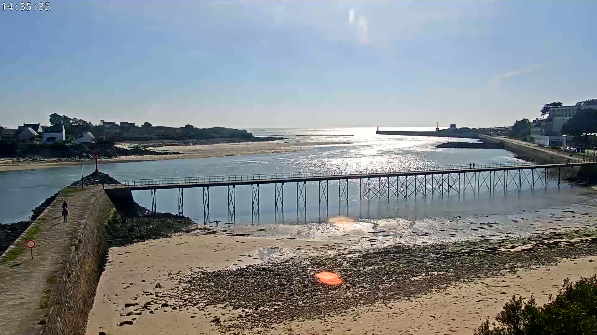 A long pier stretches across a calm, sunlit body of water, connecting two shorelines with buildings and sandy beaches visible in the background under a clear sky.