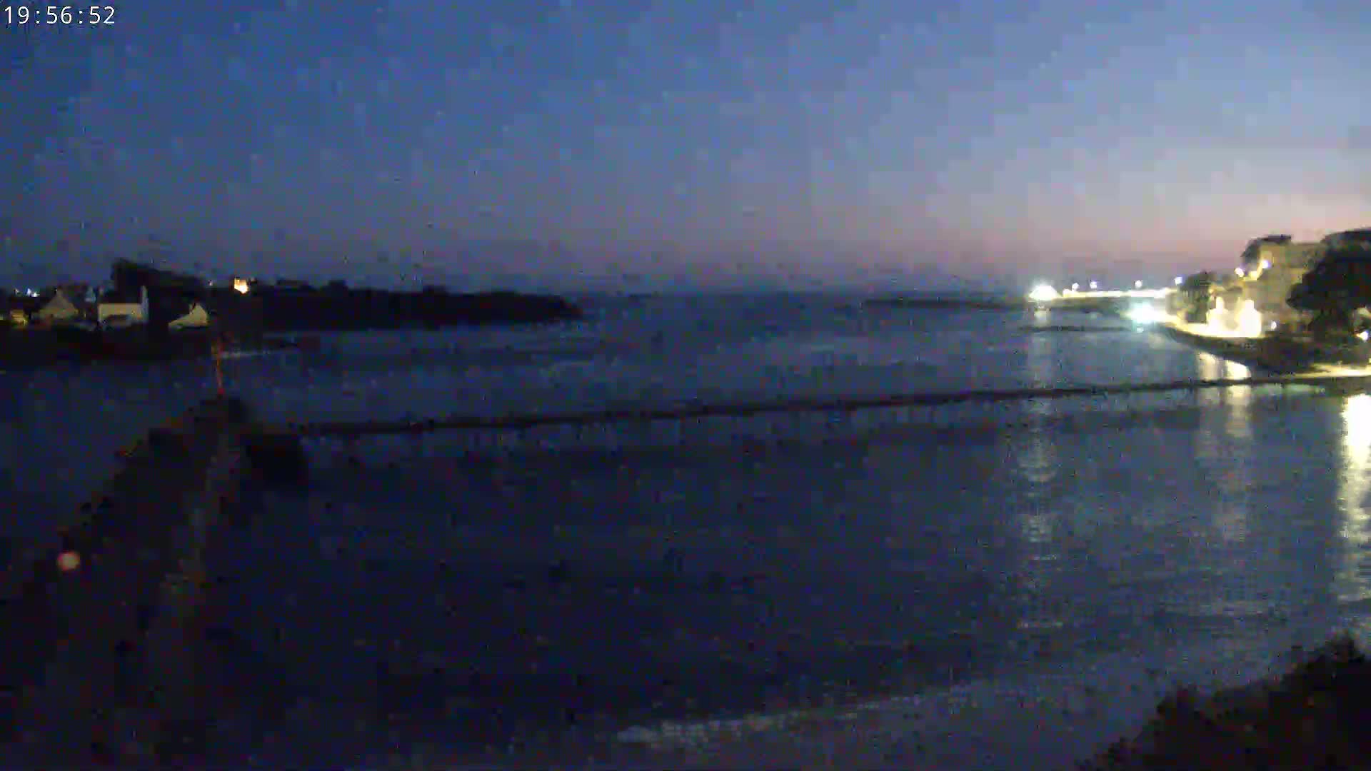 A dark, calm body of water is seen at twilight, with a long pier stretching across it, separating buildings on one side from a small island on the other.