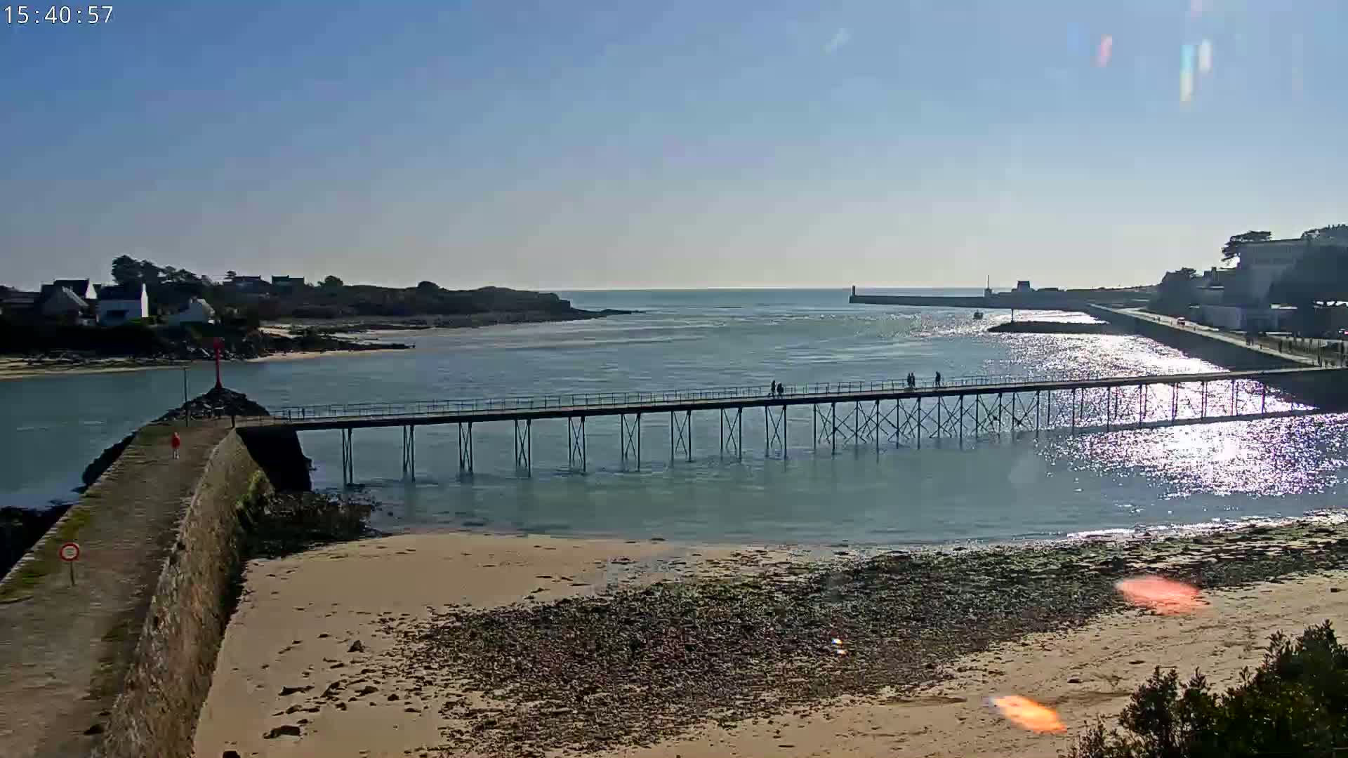 A long pier extends over calm, sunlit water toward a distant shoreline, with a sandy beach in the foreground under a clear blue sky.