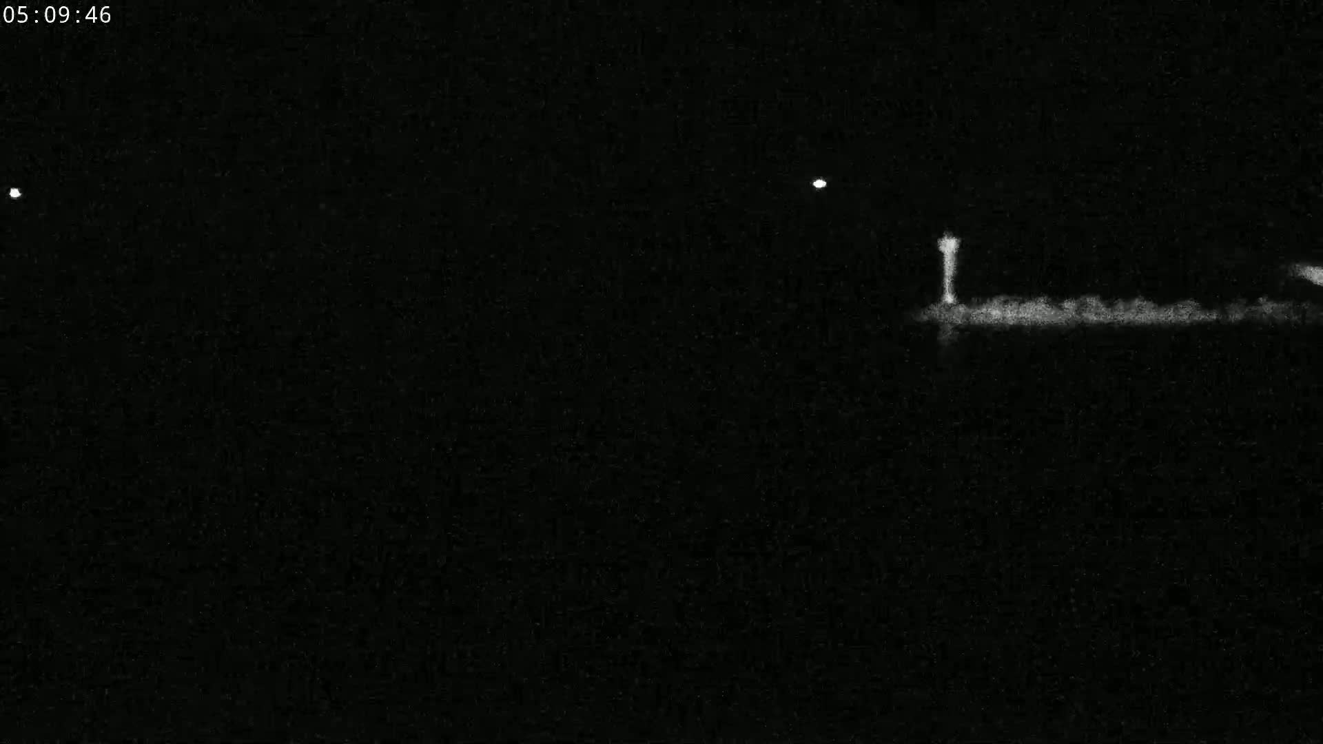 A nighttime image shows a low-lying structure, possibly a pier or shoreline, with a tall, thin light source standing in the middle against a dark, seemingly clear sky.