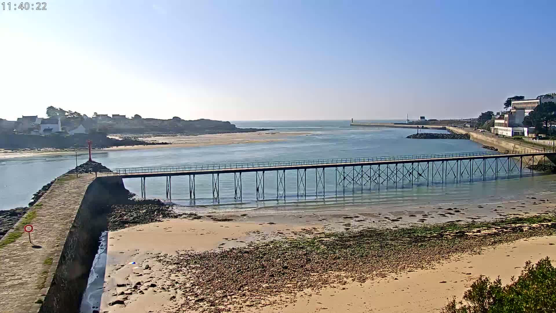 A long pier extends over calm, shallow water toward a small town nestled on a rocky point, under a clear blue sky.