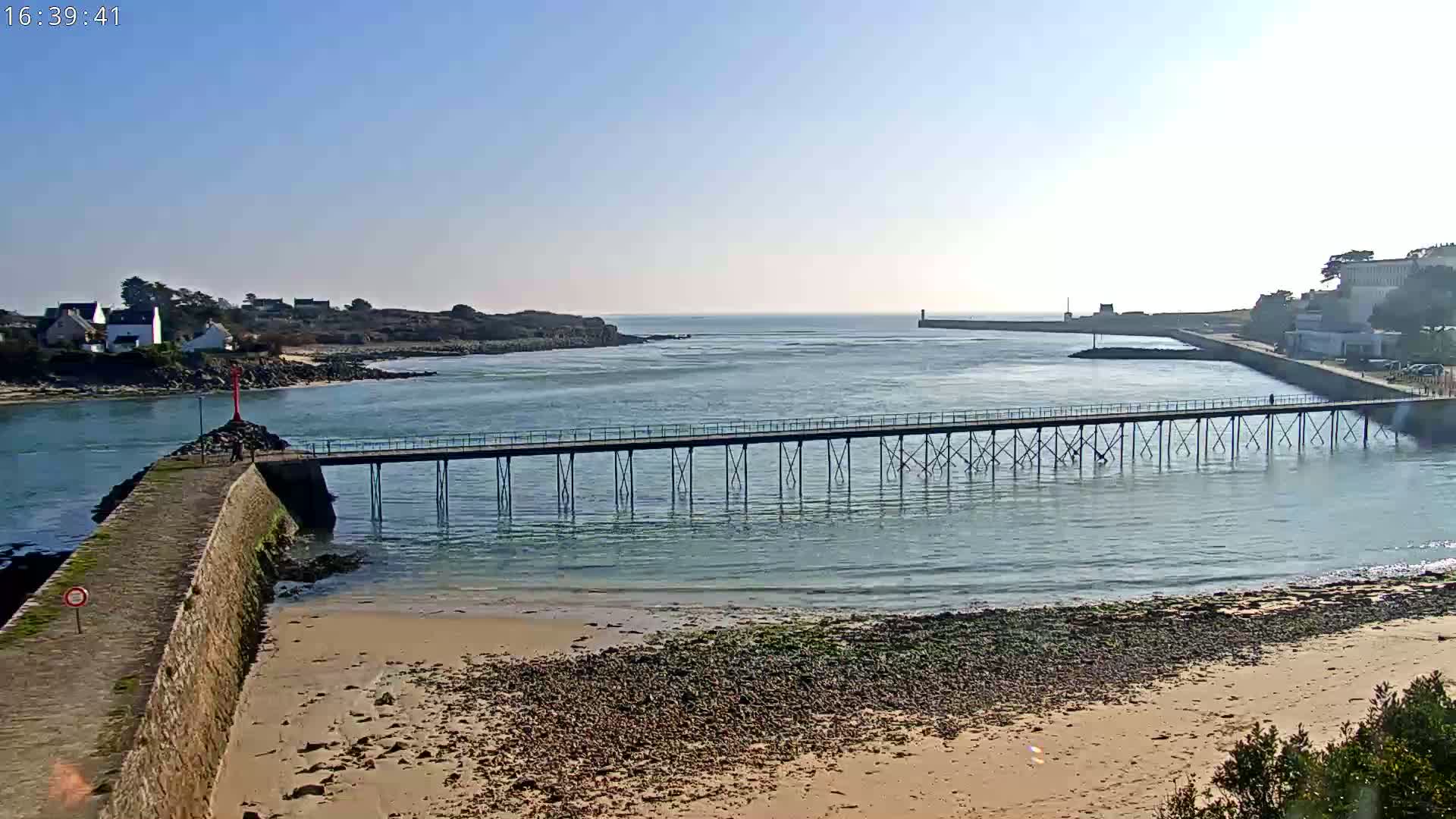 A long pier extends over calm, blue water towards a distant shoreline under a clear, bright sky, with a sandy beach in the foreground.