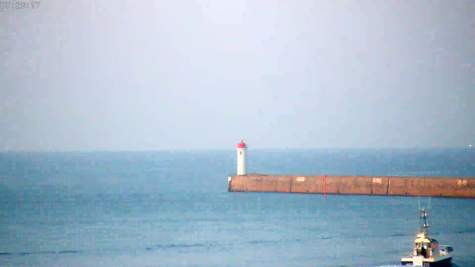 A small, white lighthouse with a red top sits atop a long, low breakwater extending into a calm, blue sea, with a small boat visible in the lower right.
