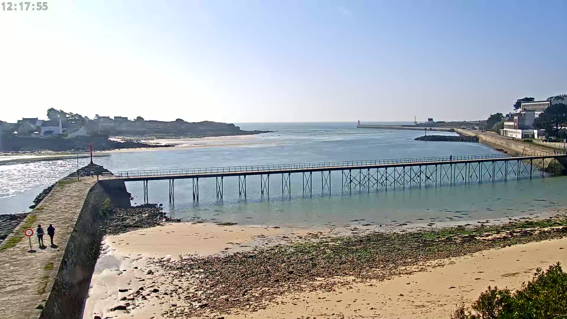 A long pier extends over calm, shallow water toward a small town and island under a clear, sunny sky.