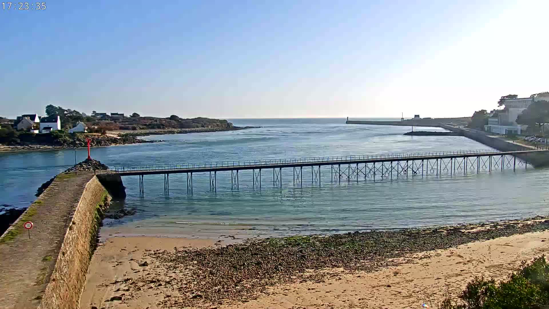 A long pier stretches across calm, light-blue water under a clear, bright sky, connecting a sandy beach to a small town and a larger landmass in the distance.