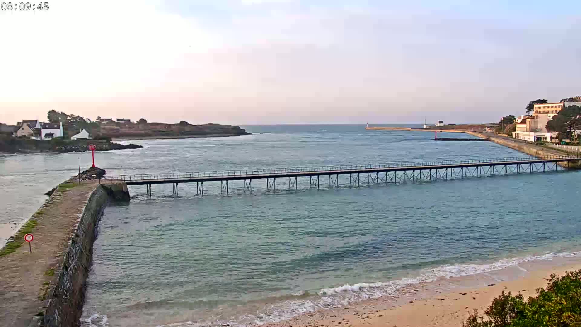 A long pier stretches across calm, light-blue water toward a distant shoreline under a pale, clear sky.