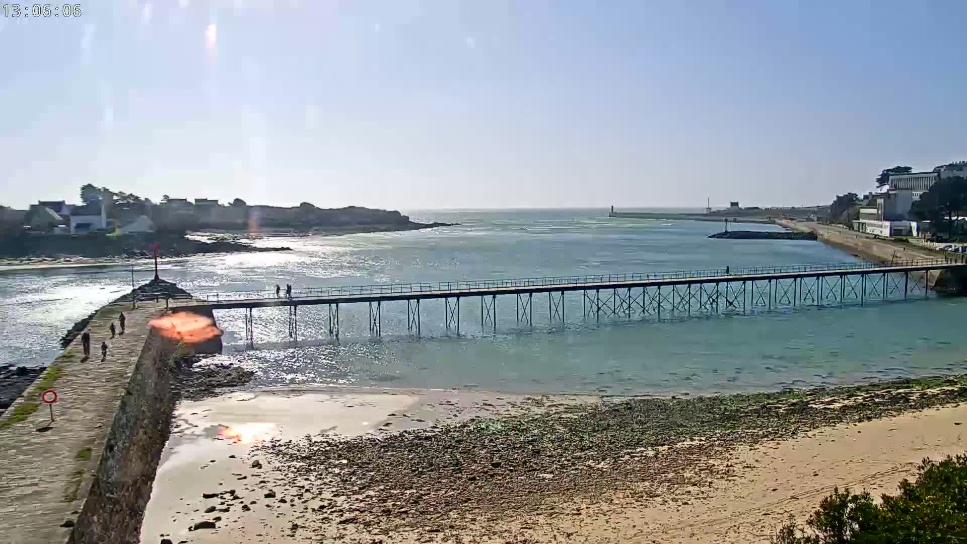 A long pier extends over calm, light-blue water toward a distant landmass under a bright, sunny sky.