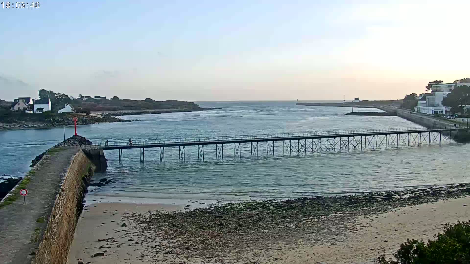 A long pier stretches across calm, shallow water separating a sandy beach from a shoreline with buildings under a clear, twilight sky.