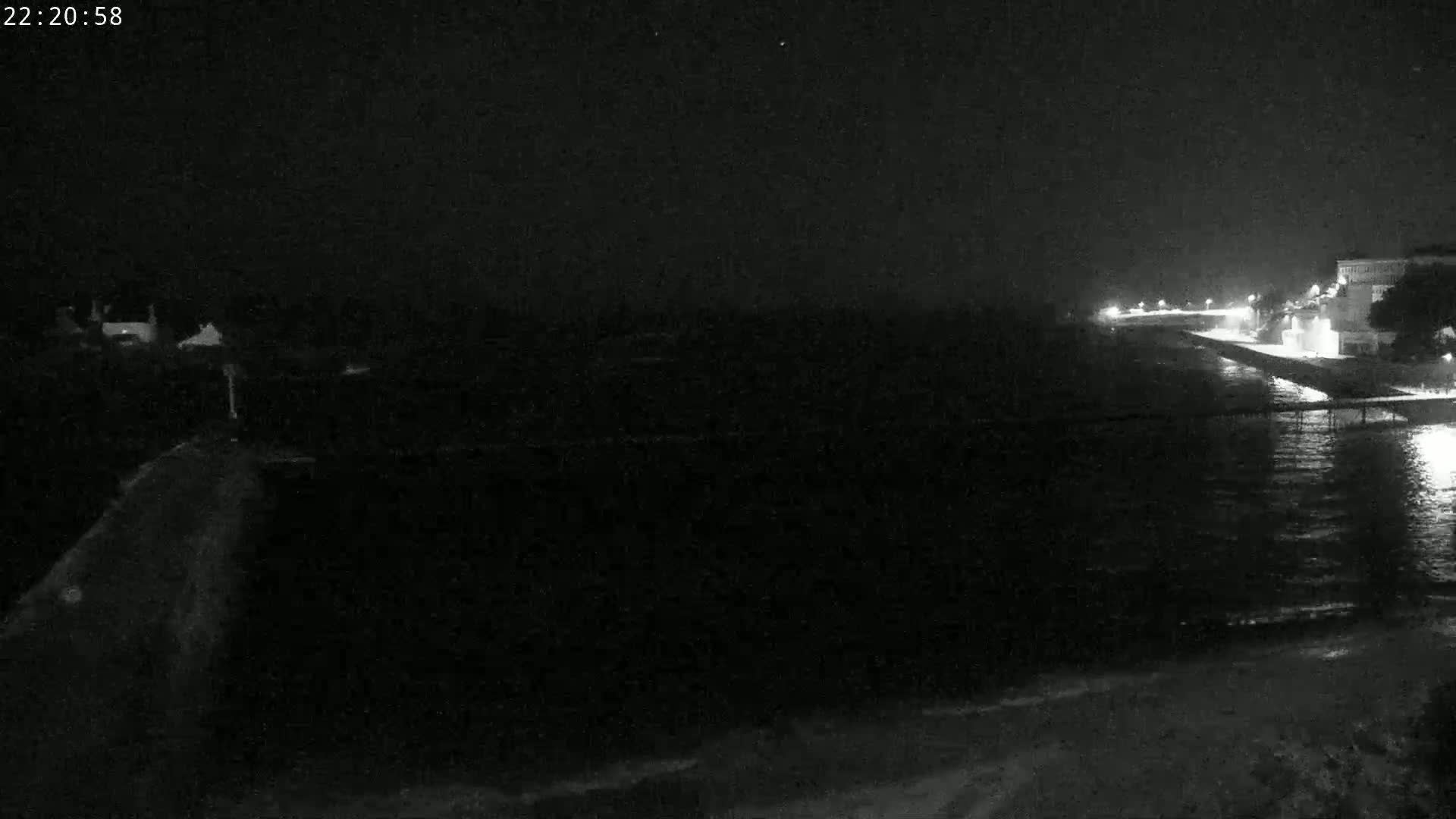 A dark, nighttime view of a calm body of water next to a dimly lit shoreline with buildings in the distance.