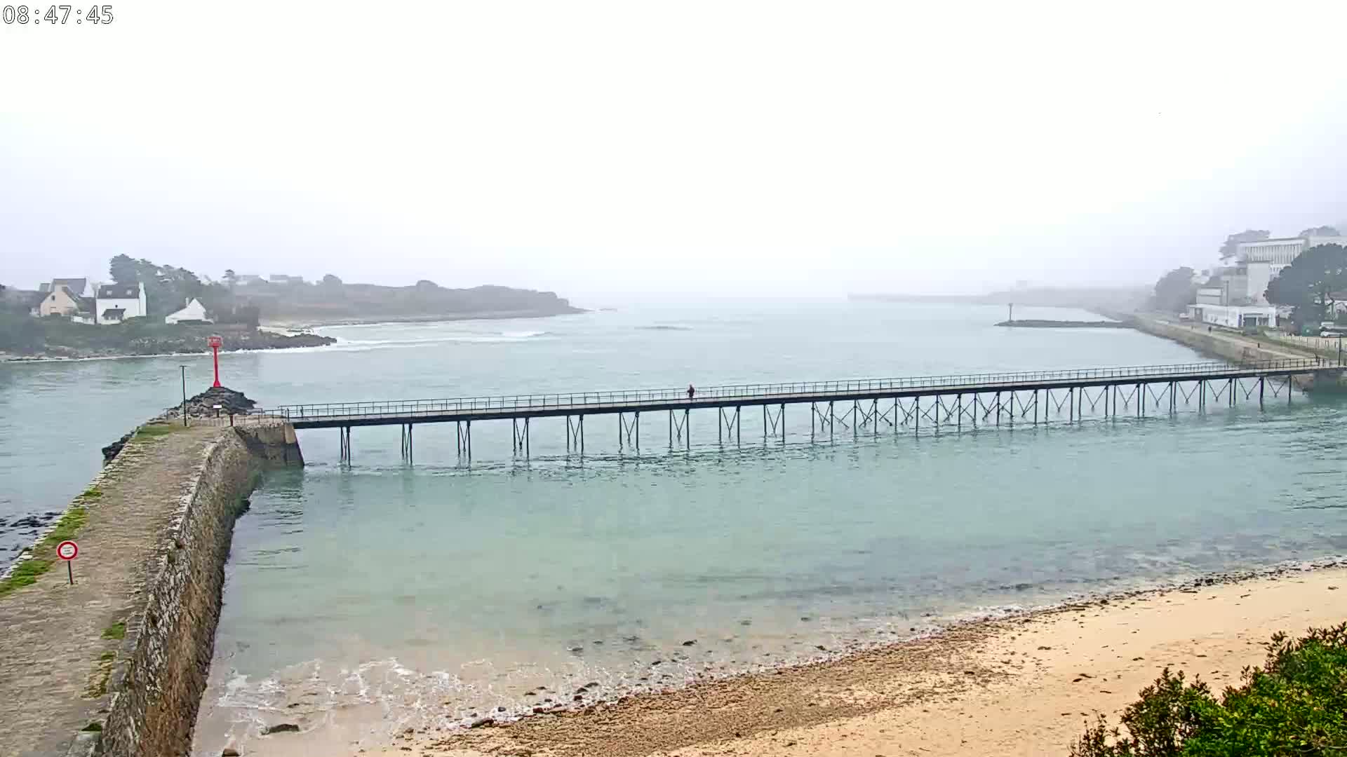 A long pier extends across calm, light-green water towards a foggy coastline with buildings and small hills visible in the distance.