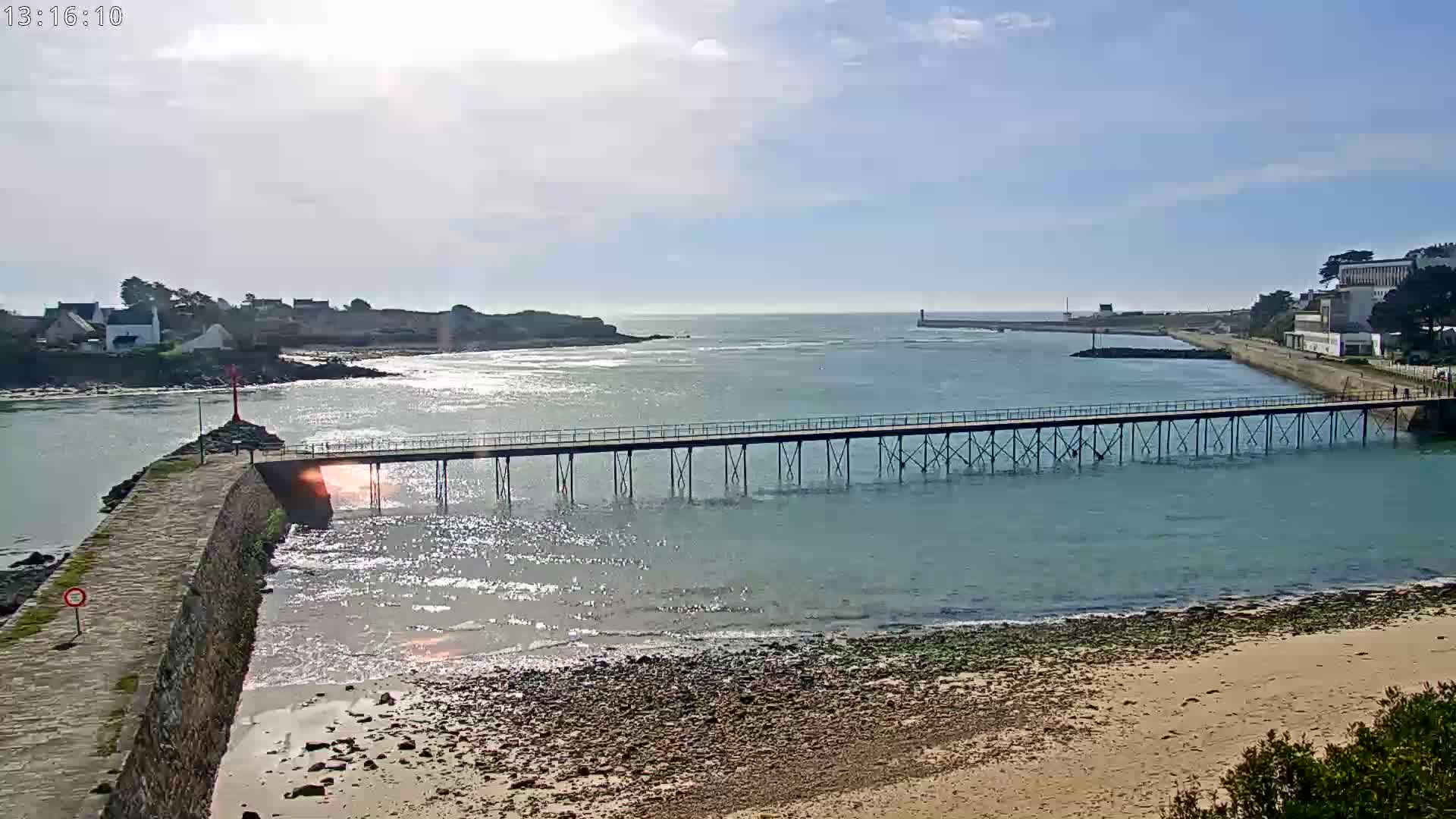 A long pier extends across calm, sunlit water toward a small island and distant shoreline under a mostly sunny sky.