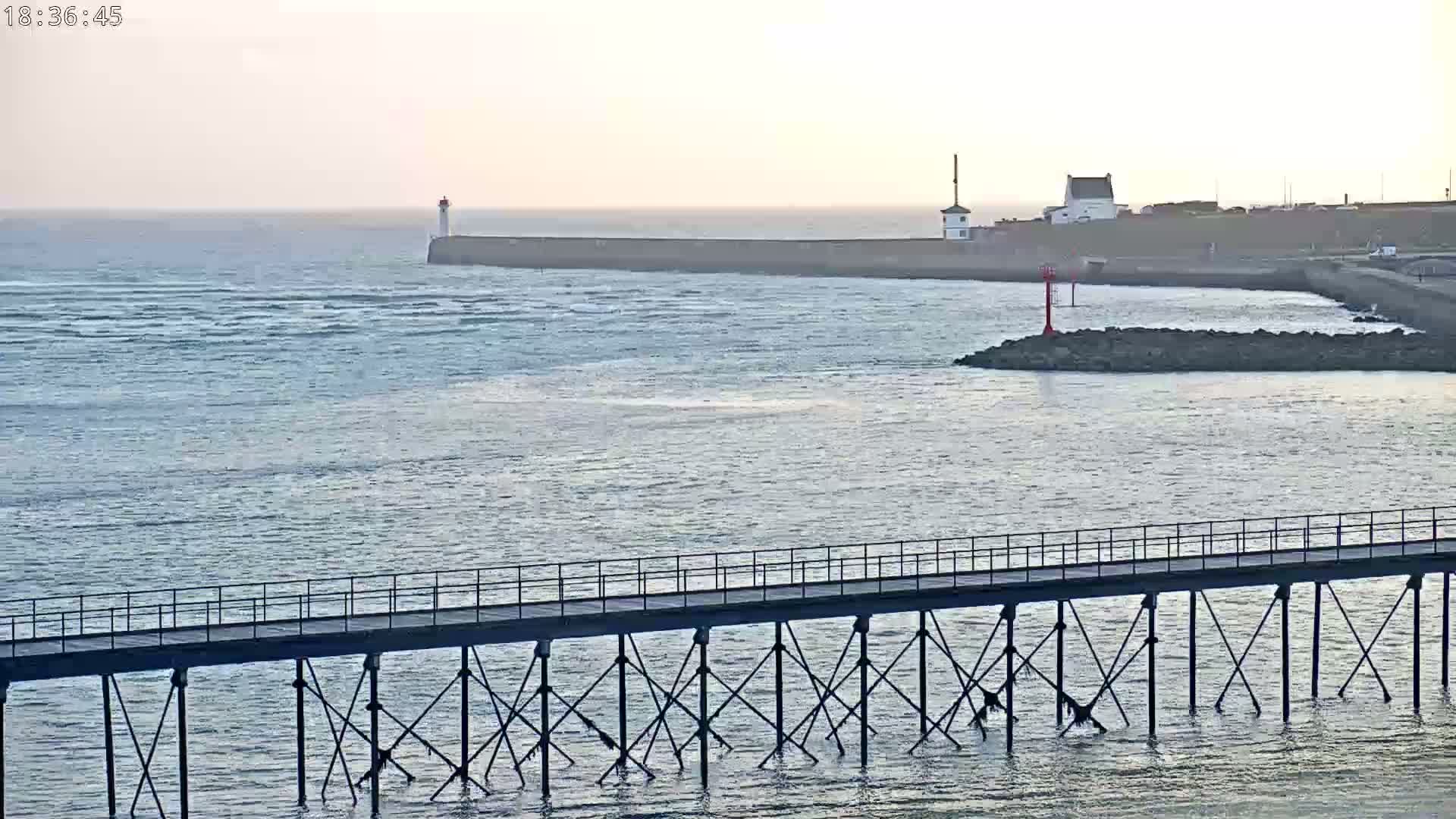 A long pier extends over a calm body of water towards a breakwater with a lighthouse and a small building, under a pale sky.