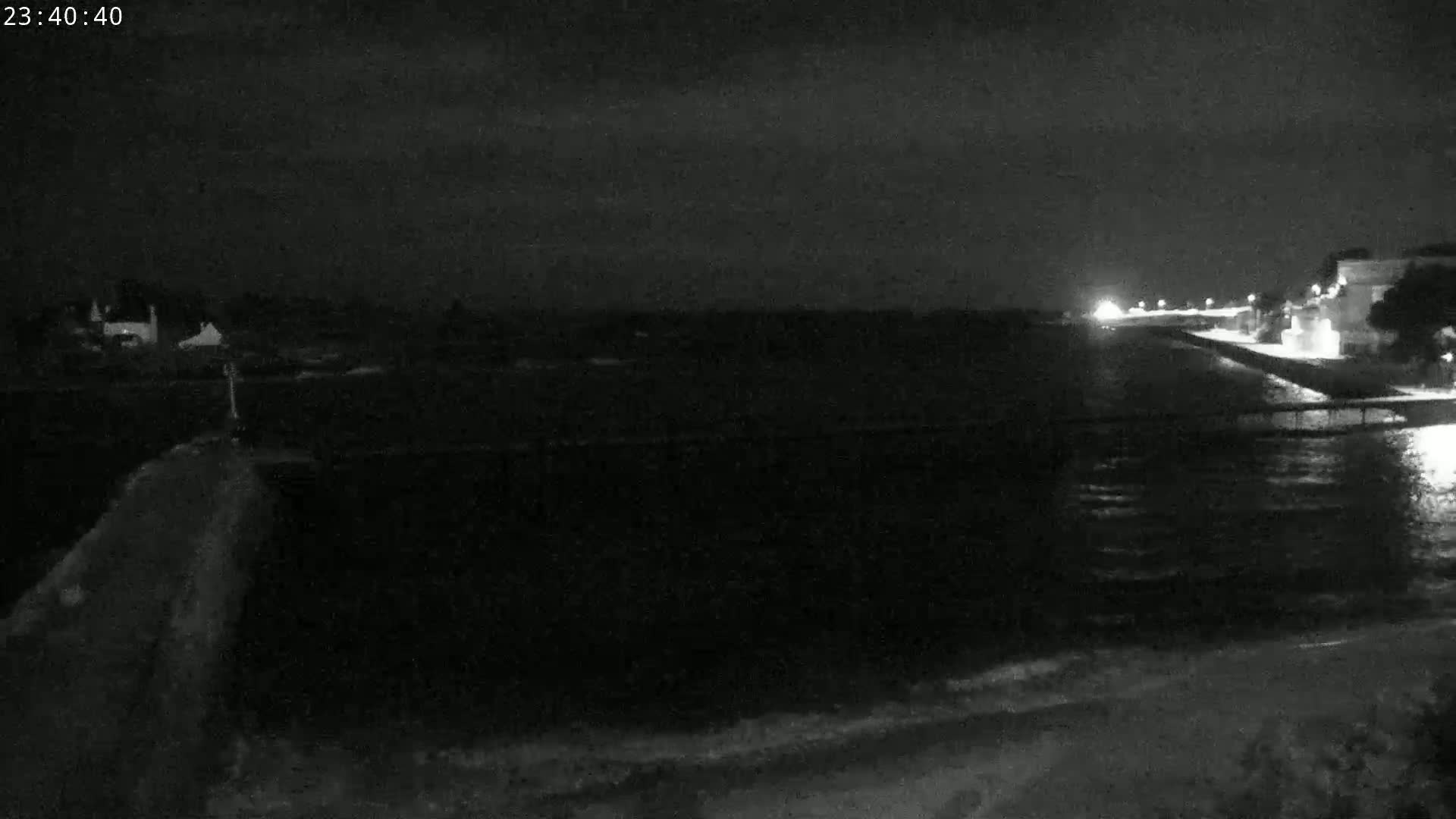 A nighttime, low-light view of a dark body of water with a shoreline featuring buildings and a seawall.