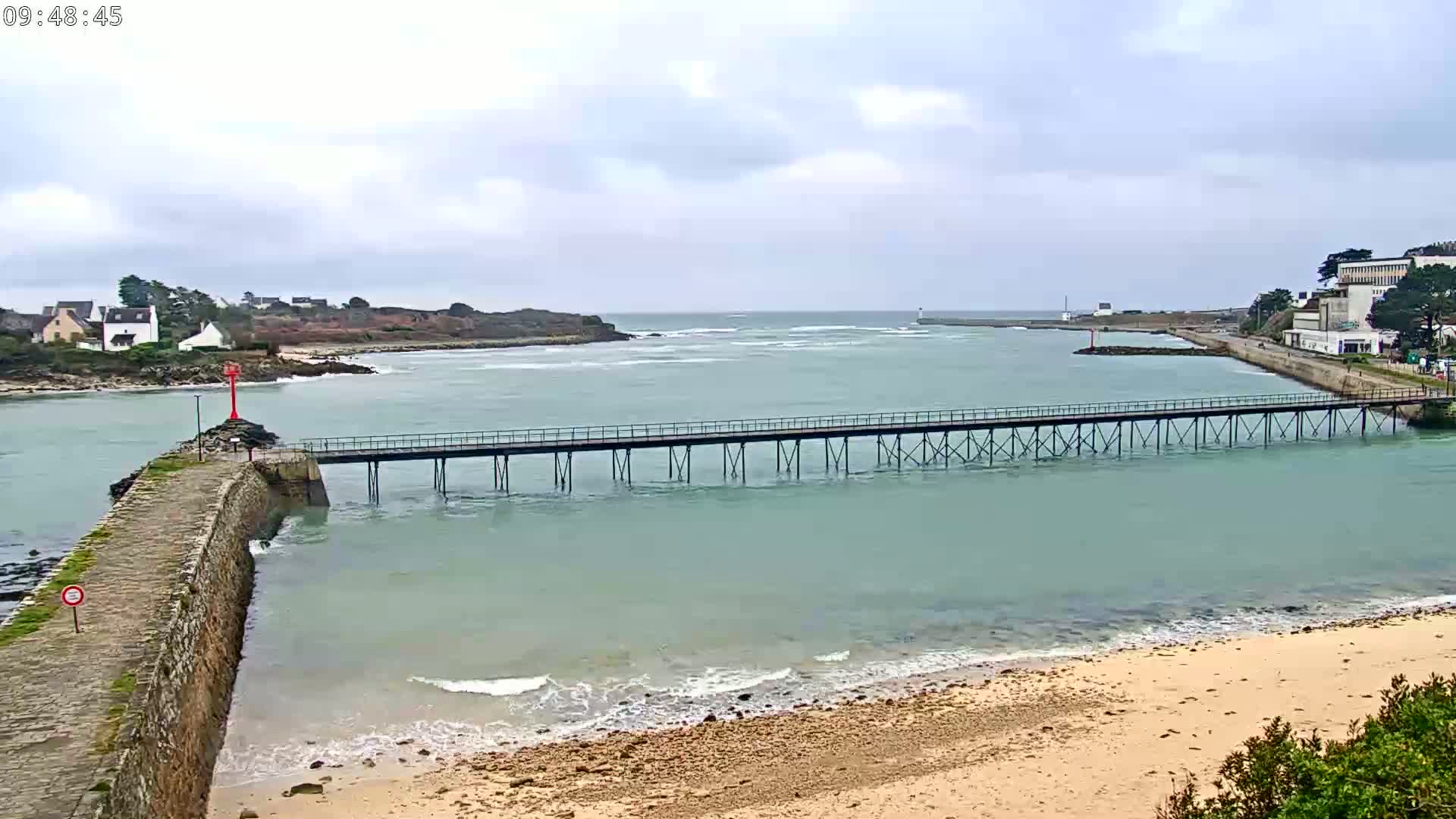 A long pier stretches across calm, light-green water toward a distant shoreline under an overcast sky, with a sandy beach in the foreground.