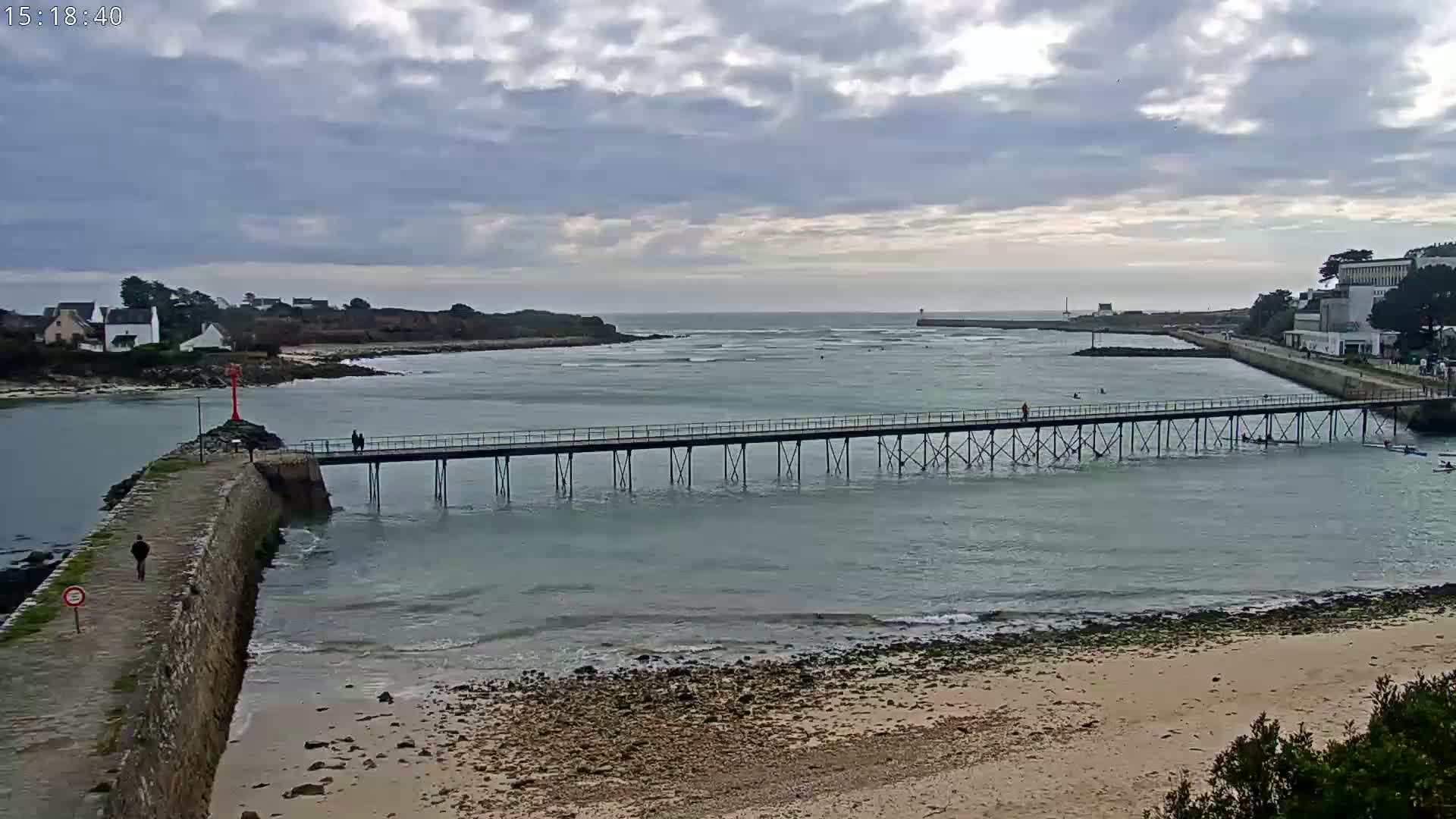 A long pier extends over calm, light-green water towards a small town under a mostly cloudy sky.