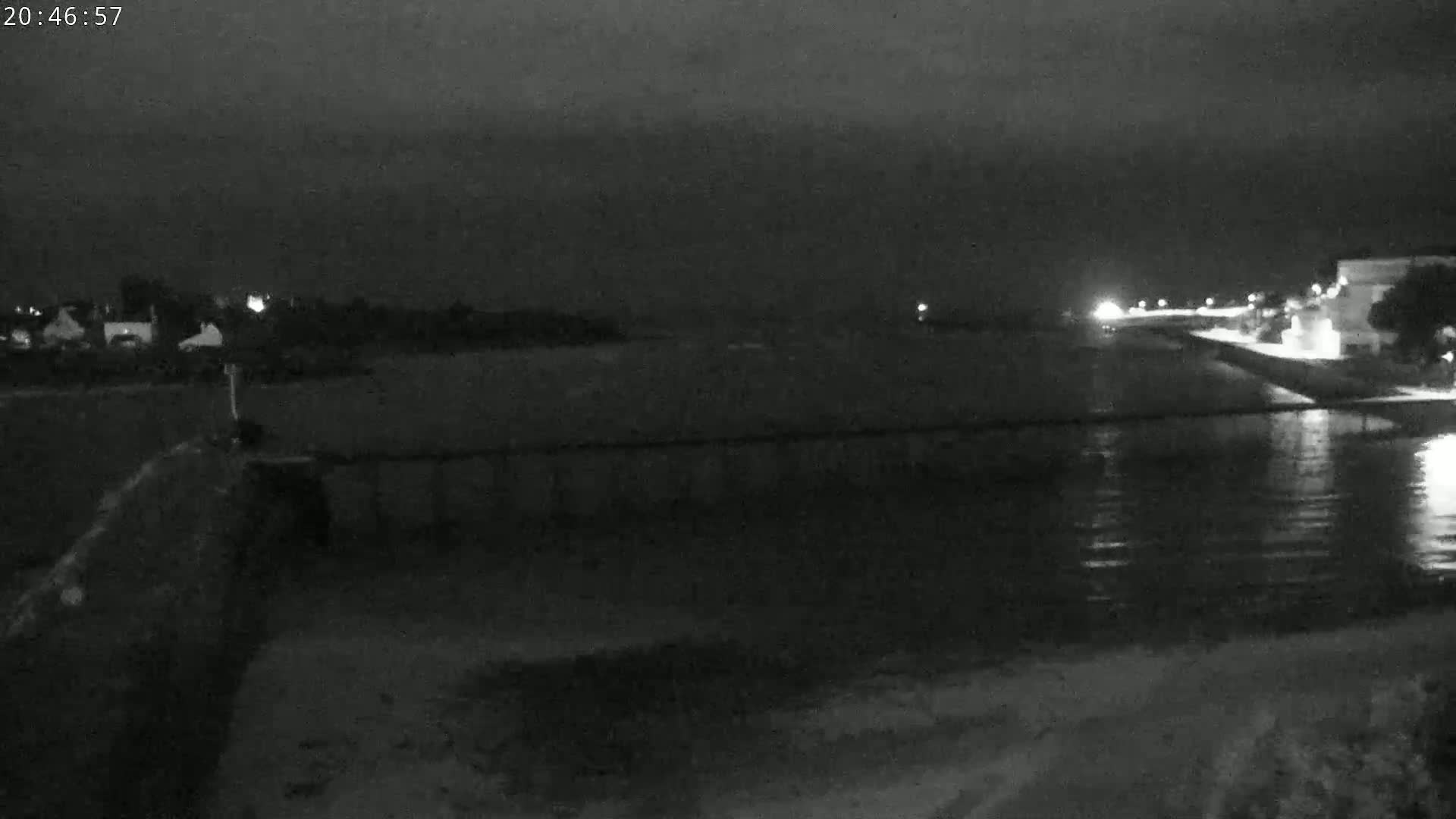 A nighttime, low-light view of a calm body of water next to a shoreline with buildings and trees visible in the distance.
