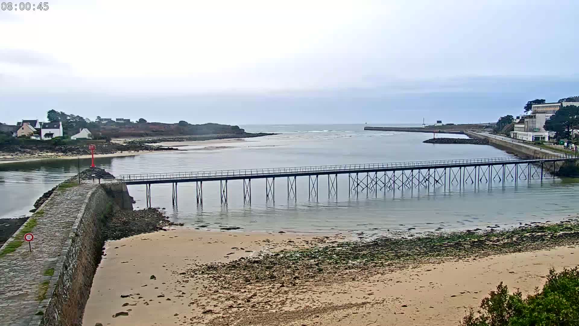 A long pier extends across a calm body of water under an overcast sky, with sandy shores and buildings visible in the distance.