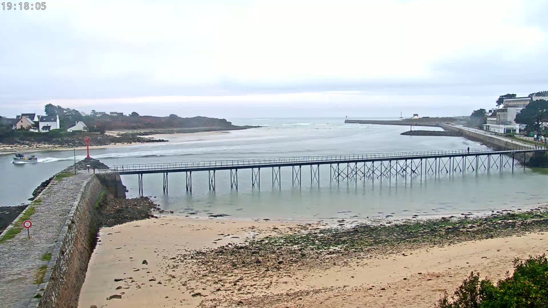 A long pier extends over calm, shallow water toward a small town and distant island under an overcast sky.