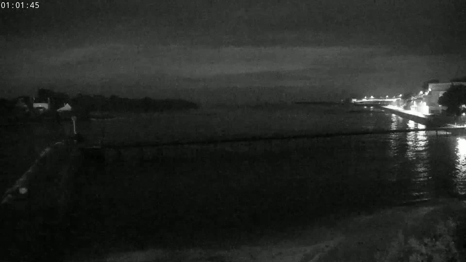 A nighttime, low-light view of a calm body of water with a shoreline featuring buildings and some vegetation.