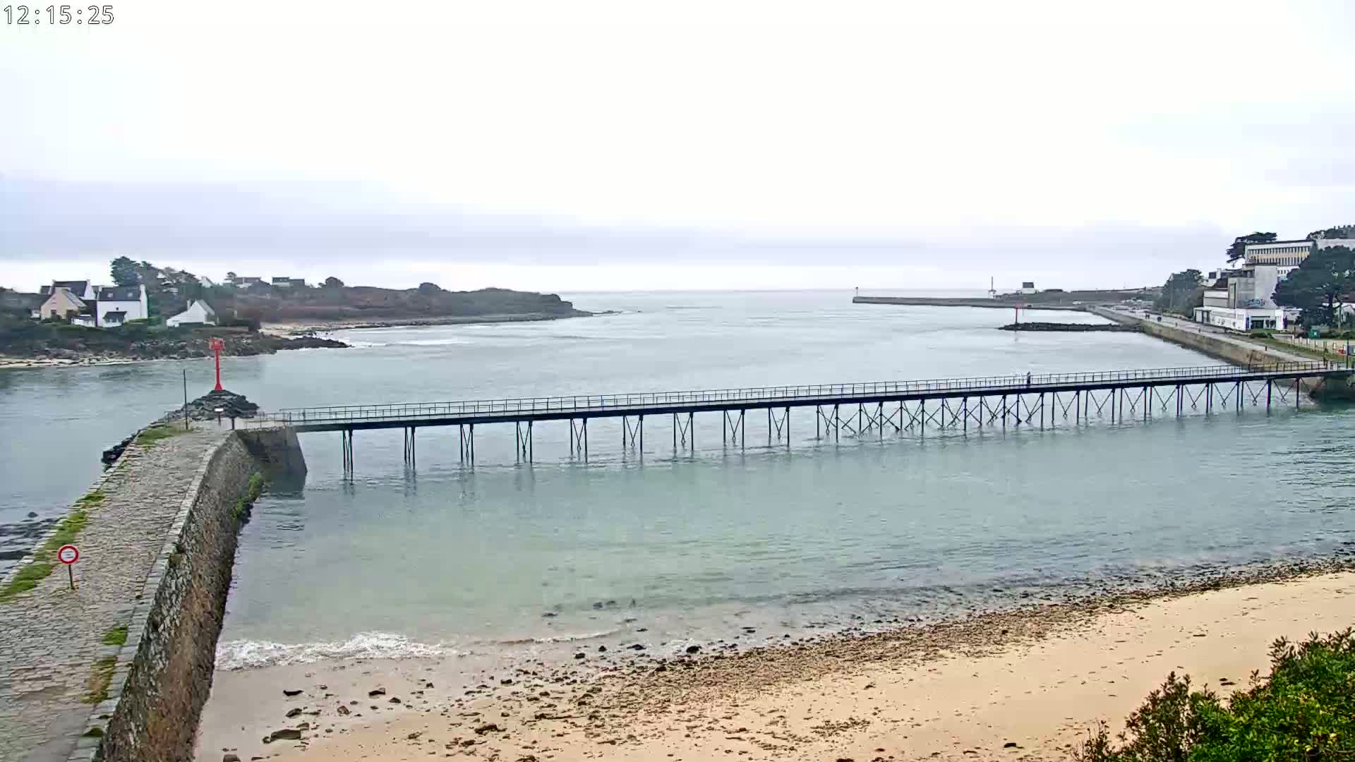 A long pier stretches across calm, light-blue water under an overcast sky, connecting a stone jetty to a shoreline with a sandy beach.
