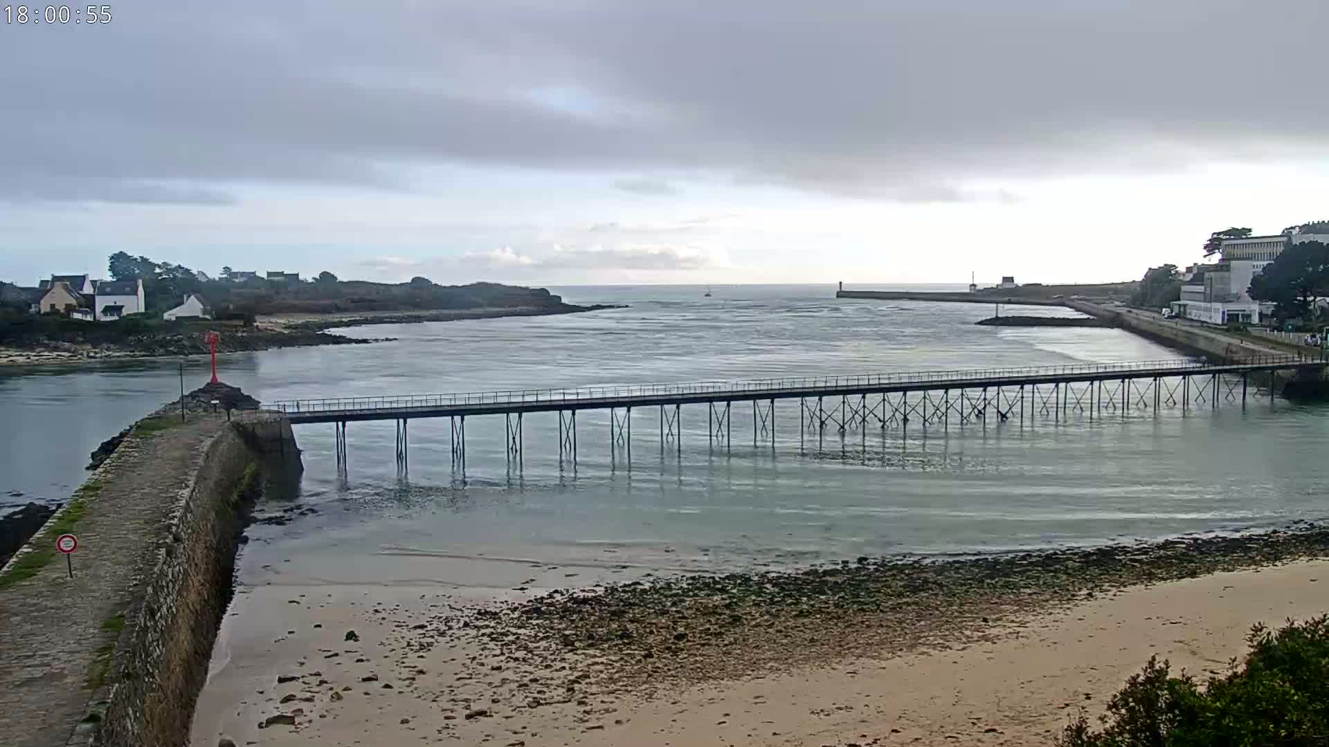 A long pier extends over calm, shallow water toward a small town under a cloudy sky.