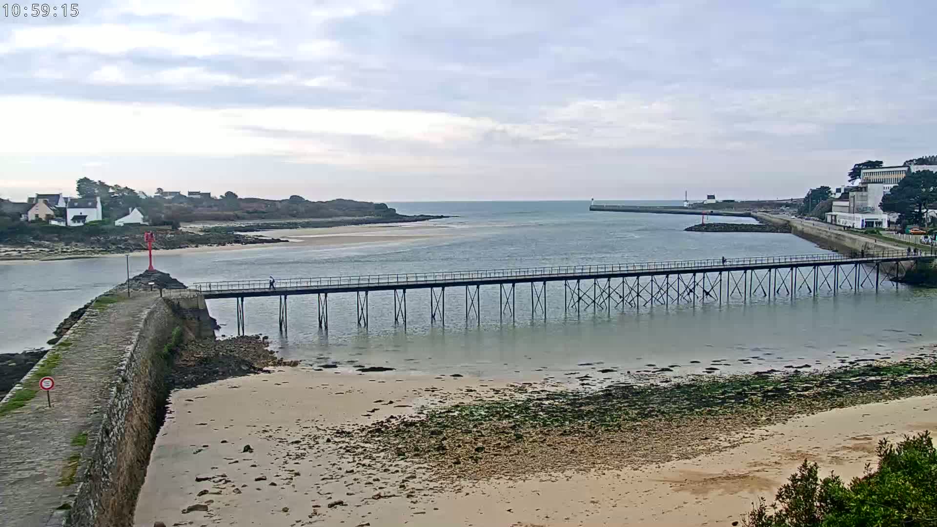 A long pier stretches across a calm, shallow waterway toward a small town and island under a cloudy sky.