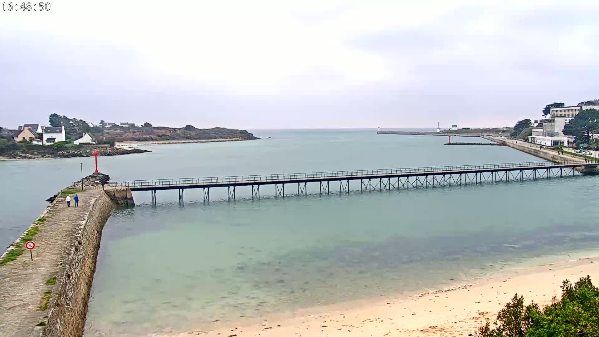 A long pier stretches across calm, light-green water toward a small island and a distant town under an overcast sky.