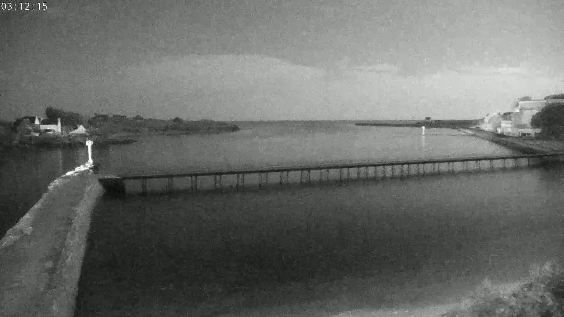A long pier stretches across a calm body of water under an overcast sky, with buildings visible on either shore.