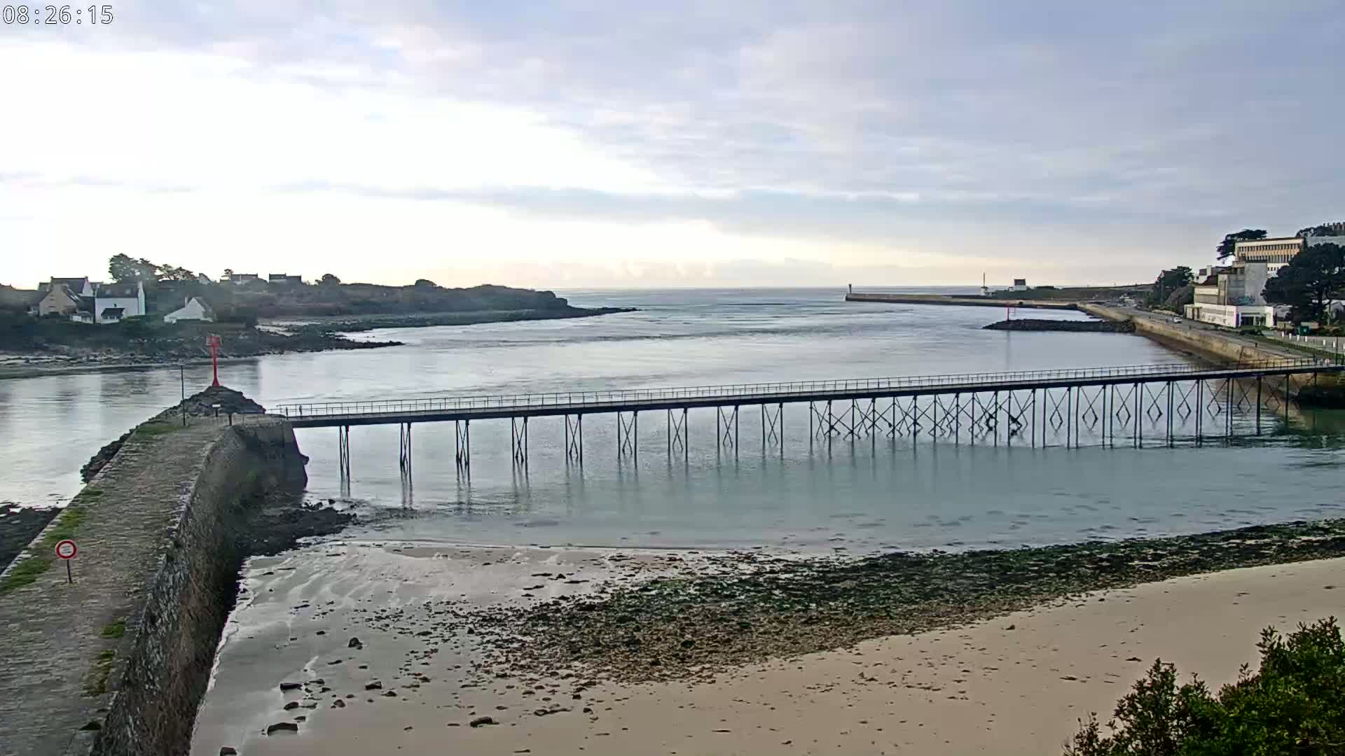 A long pier stretches across calm, shallow water toward a small town and a larger building on the opposite shore under an overcast sky.