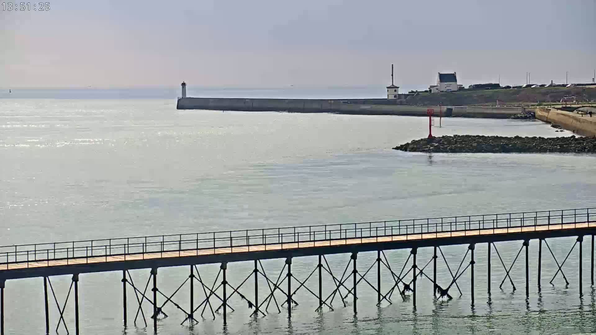 A long pier extends over calm, light-grey water toward a breakwater with a lighthouse and small building, under a hazy sky.
