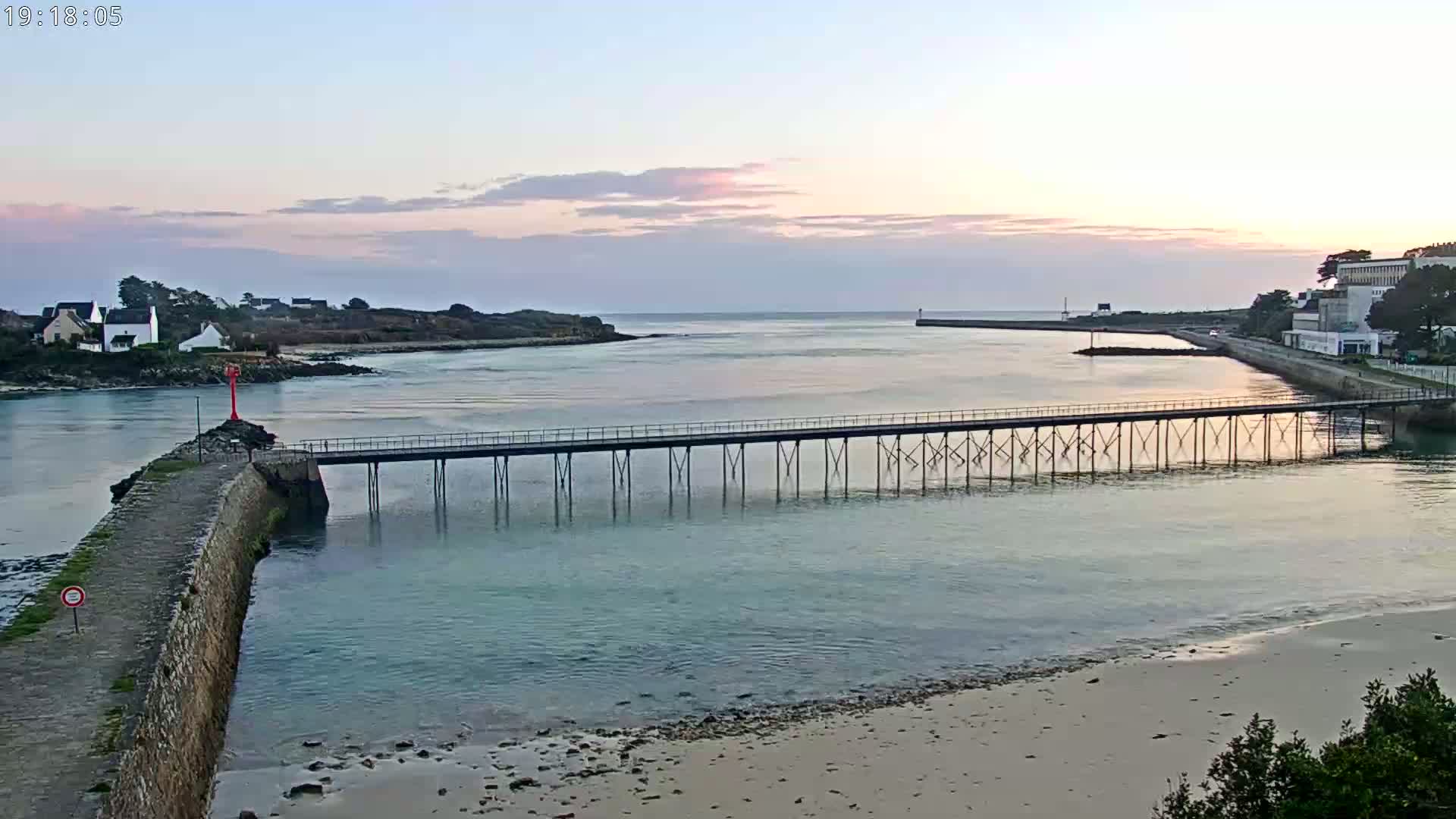 A long pier stretches across calm, light-blue water at sunset, connecting a stone wall to a shoreline with a sandy beach and buildings in the distance.
