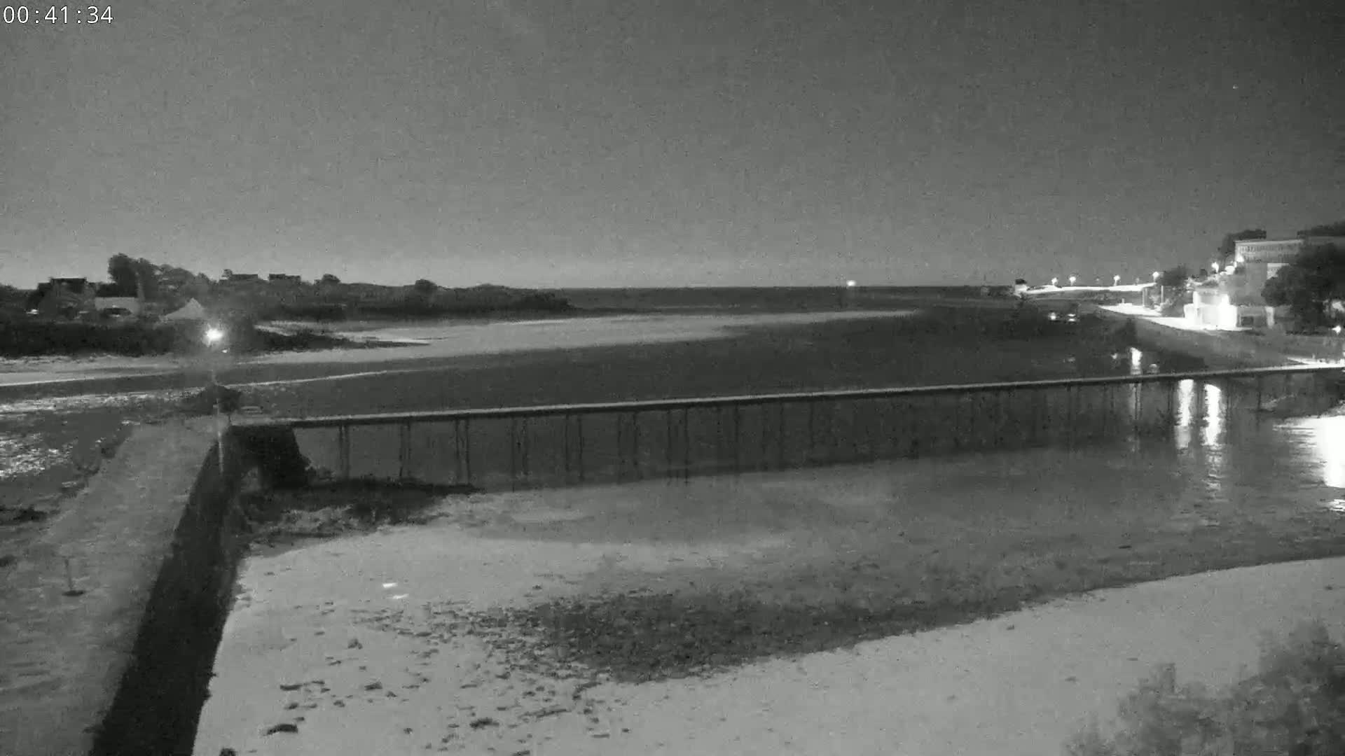 A long, low pier stretches across a calm body of water at night, with buildings visible in the distance under a dark sky.