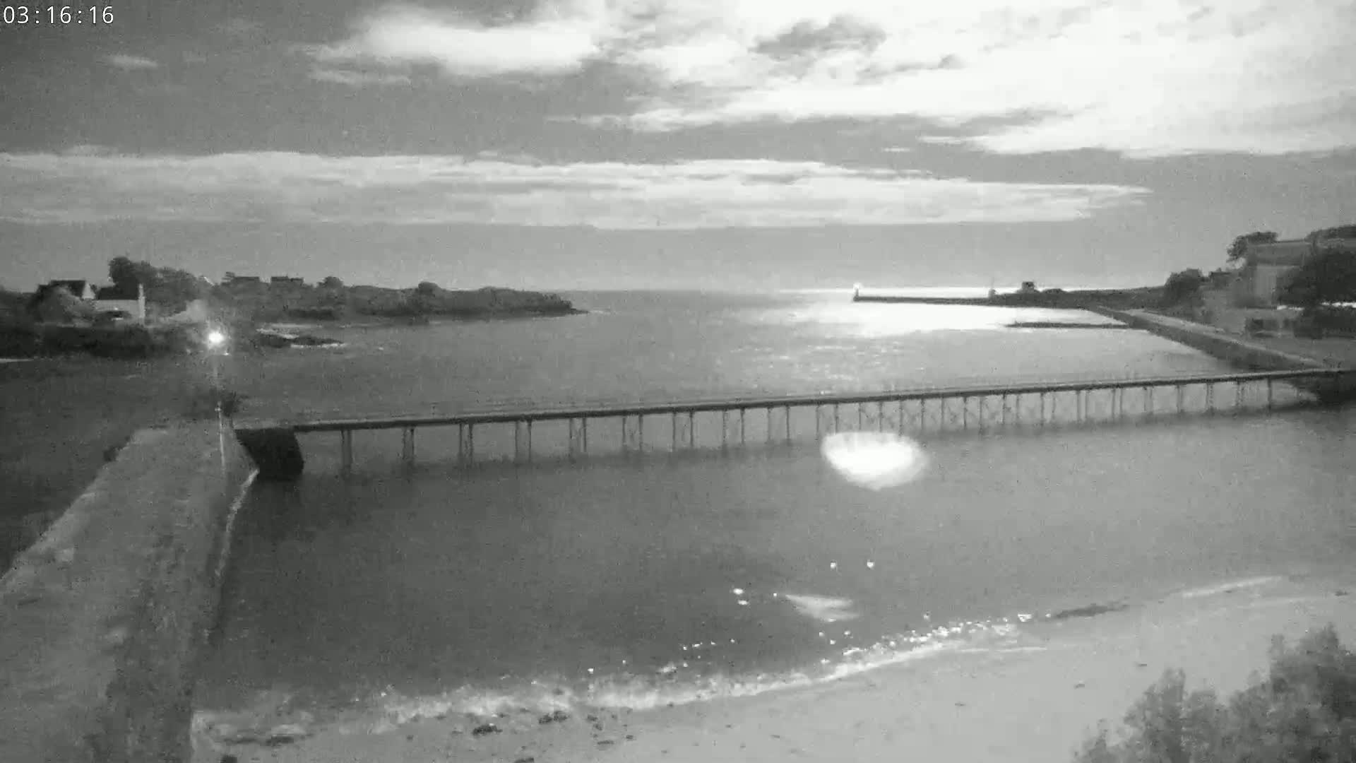 A long pier stretches across calm water under a partly cloudy night sky, with land and buildings visible on either side.