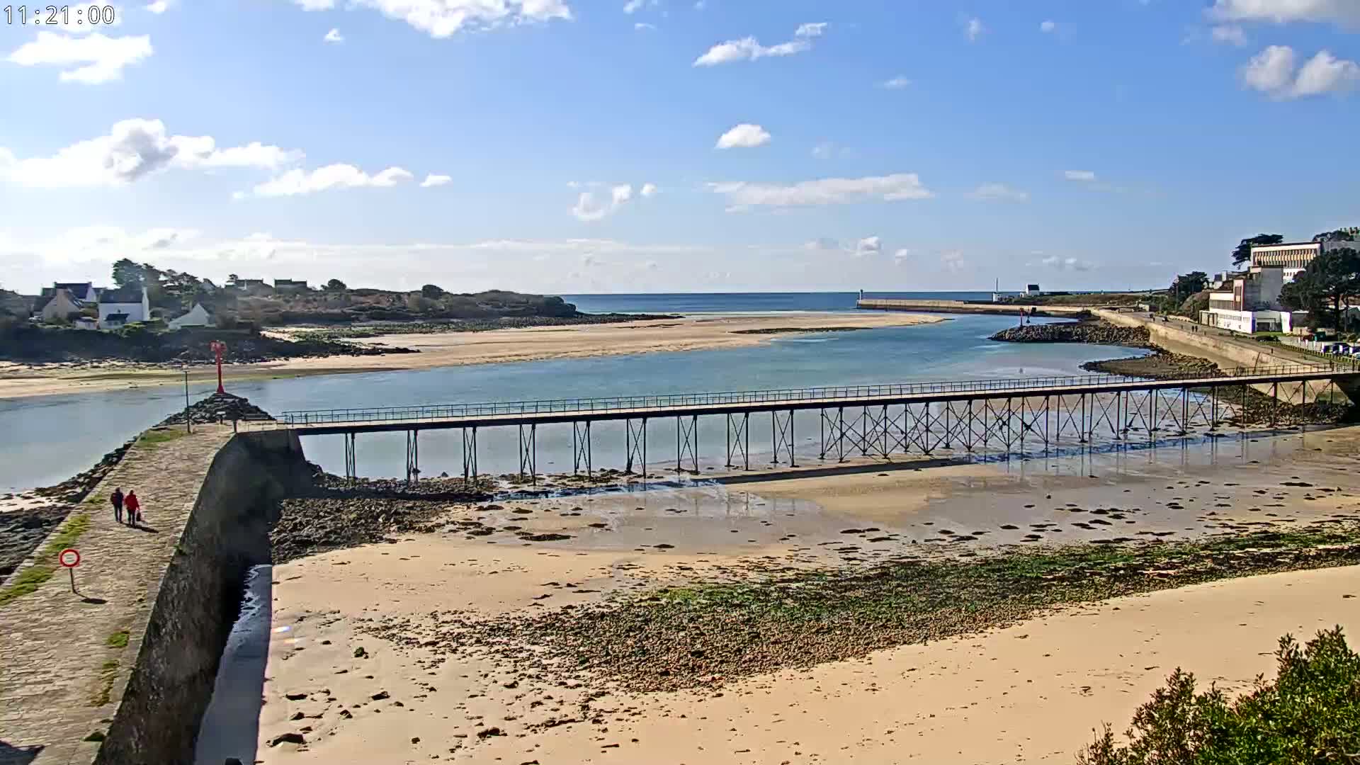 A long pier stretches across a calm, shallow waterway between a sandy beach and a small island under a partly cloudy sky.