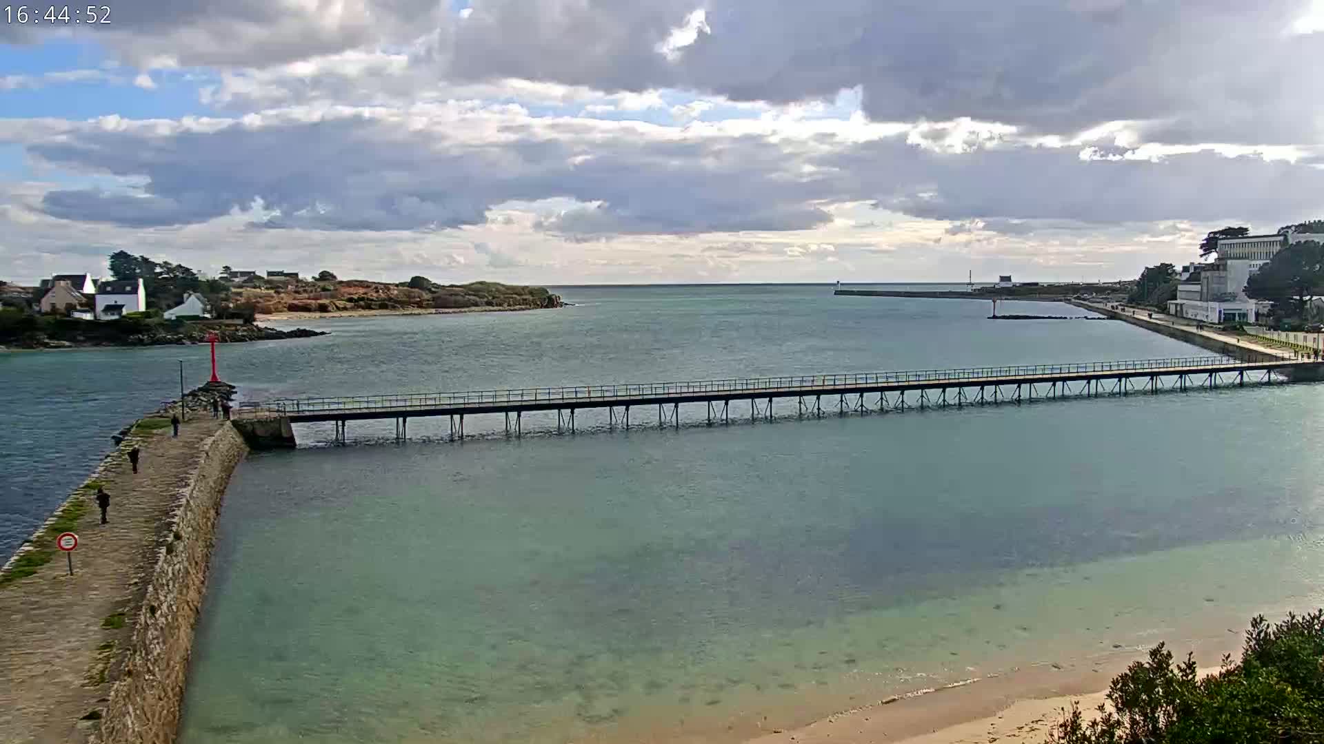 A long pier extends over calm, teal water toward a small island under a partly cloudy sky.