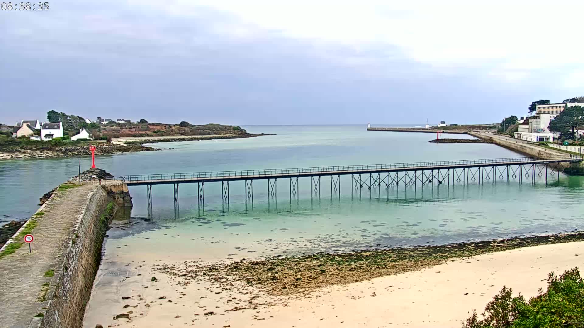 A long pier stretches across calm, shallow, teal water toward a small town and island under an overcast sky.