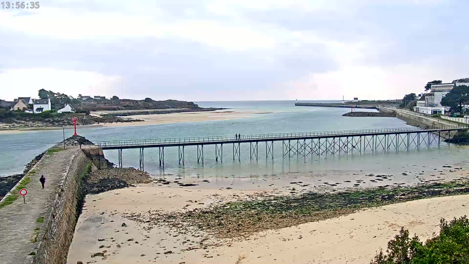 A long pier stretches across shallow, calm water toward a small island under a mostly cloudy sky.